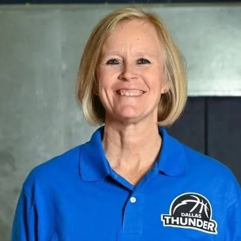A smiling woman with shoulder-length blonde hair, wearing a blue Dallas Thunder polo shirt, standing indoors against a dark and plain background.