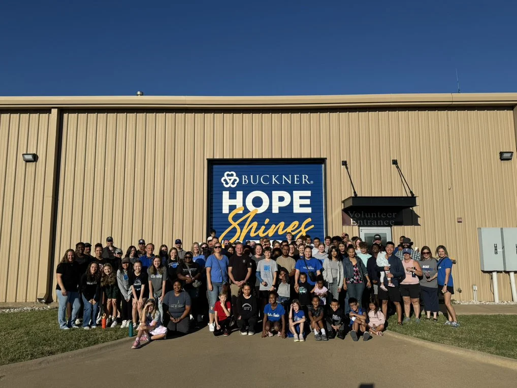 A large group of people standing outside a beige industrial building with a blue sign that reads 'BUCKNER HOPE Shines'. The group includes children and adults, some wearing blue shirts, under a clear blue sky.