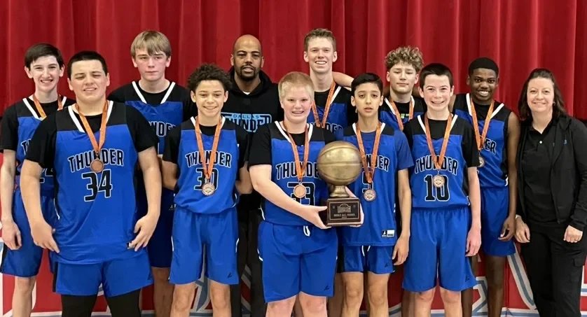 A basketball team of young boys posing with their coach, all wearing blue jerseys with 'Thunder' and medals around their necks, holding a trophy, standing in front of a red curtain.