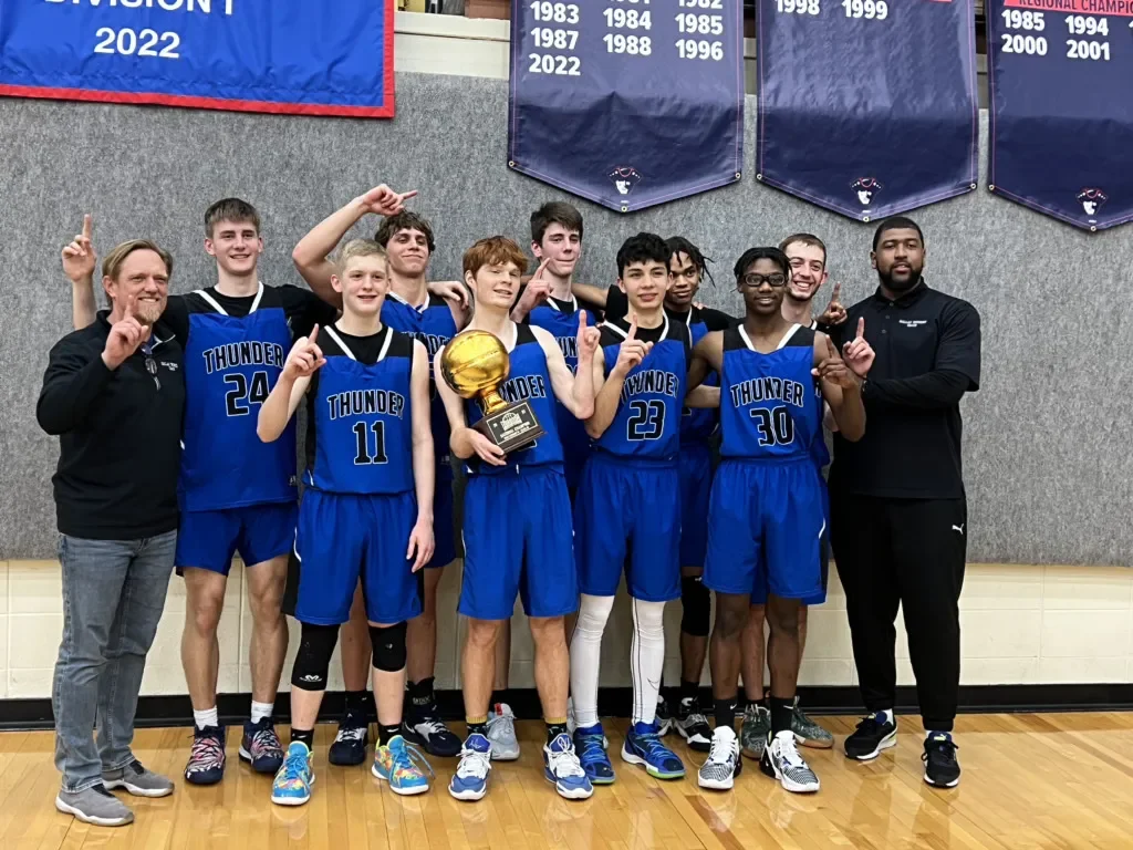 Basketball team of young boys and their coaches celebrating with a trophy and raising their hands in victory in a gymnasium.