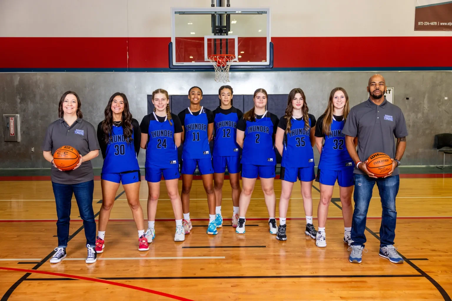 A girls' basketball team and coaches posing for a team photo in a gymnasium, with women in matching blue uniforms and two coaches holding basketballs, standing in front of a basketball hoop.