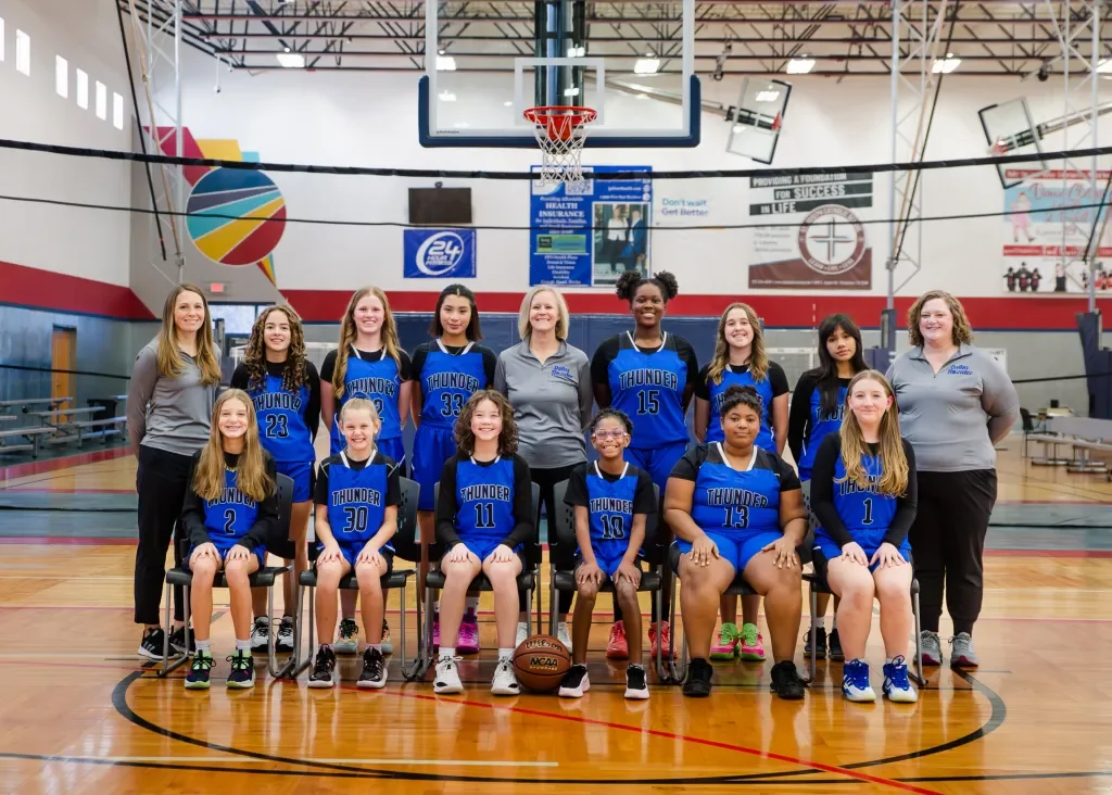 A girls' basketball team called the Thunder posing for a team photo in a gymnasium, with coaches standing on either side and basketball court in the background.
