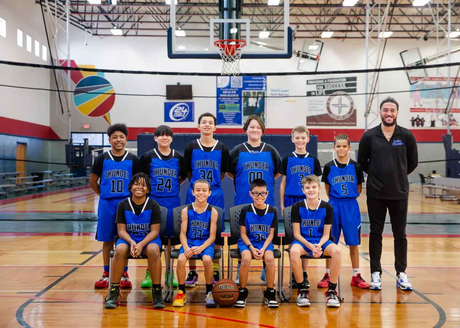 A youth basketball team called the Thunder with a coach posing in an indoor gymnasium. The team consists of nine children in blue and black uniforms and one adult coach standing at the right end. The children are seated and standing in two rows in front of the basketball hoop.