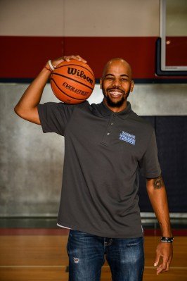 A man standing in a basketball gym, smiling, holding a basketball over his shoulder.
