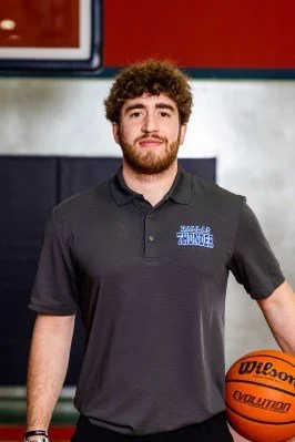 A young man with curly hair and a beard standing in a gym, holding a basketball, wearing a black polo shirt with a logo on the chest.