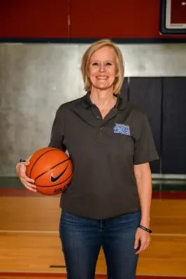 A woman standing in a basketball court holding a basketball, smiling, wearing a dark gray polo shirt and jeans