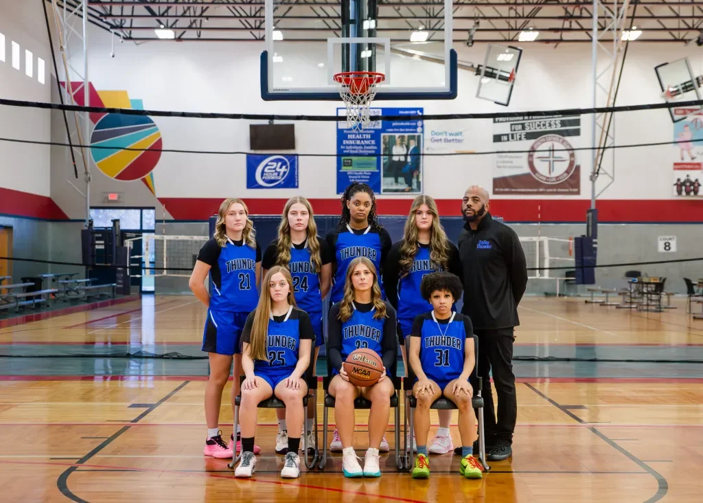 A school basketball team called the Thunder, including seven players and their coach, posing for a team photo inside a gymnasium. The players are dressed in blue jerseys with numbers and team name, with some sitting and others standing, and a basketball is held by one of the seated players.