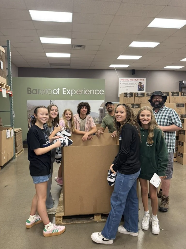 Group of eight people, mostly young girls, standing around a large cardboard box on a pallet inside a warehouse, with shelves and packing instructions in the background.