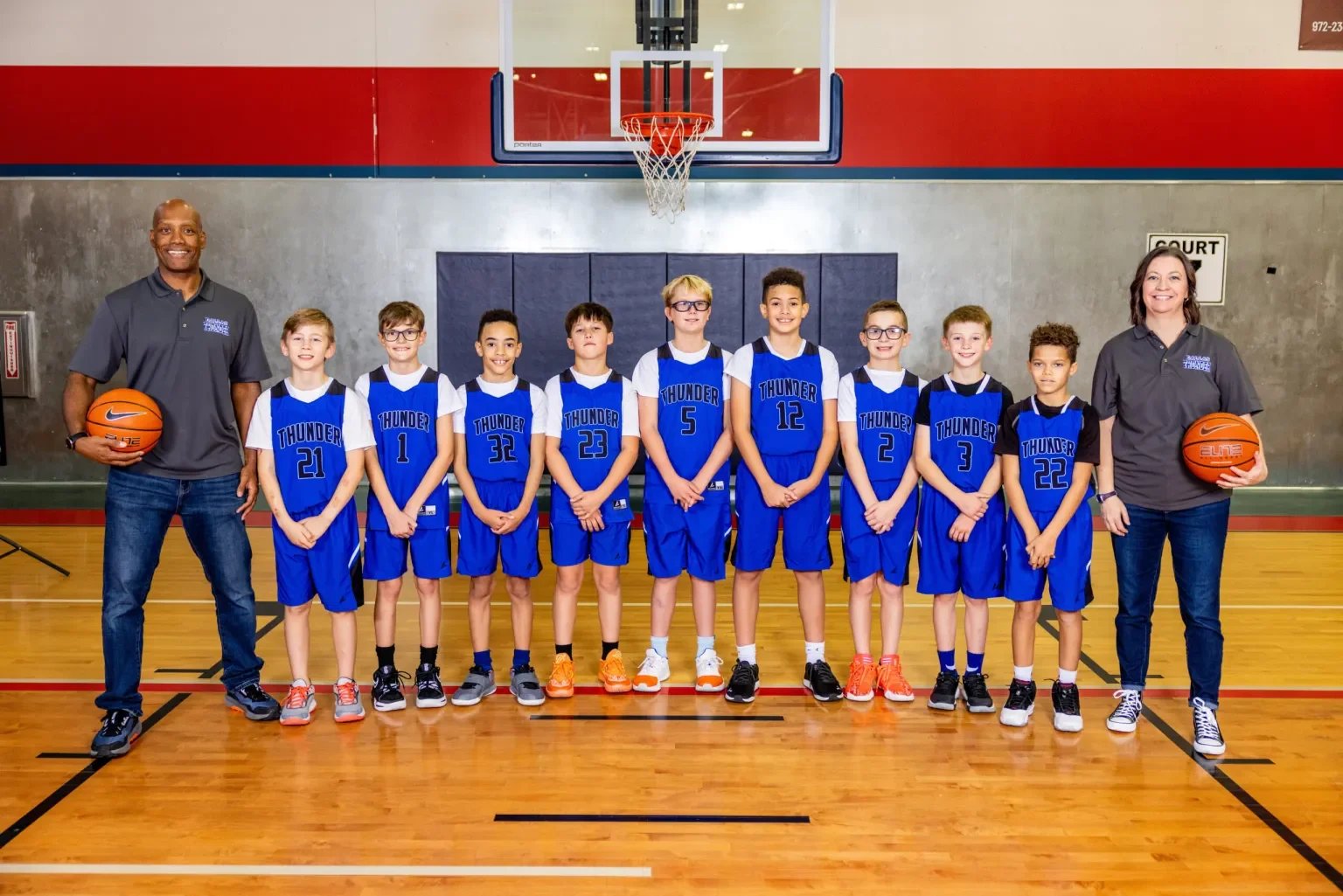 A youth basketball team called Thunder with their two coaches, a man and a woman, standing on a basketball court in a gymnasium. The players are wearing blue uniforms with their team name and numbers, and the coaches are holding basketballs.