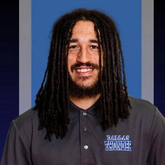 A man with long dreadlocks, a beard, and a smiling face is wearing a gray sports polo shirt with the Dallas Thunder logo on it, standing against a blue background.