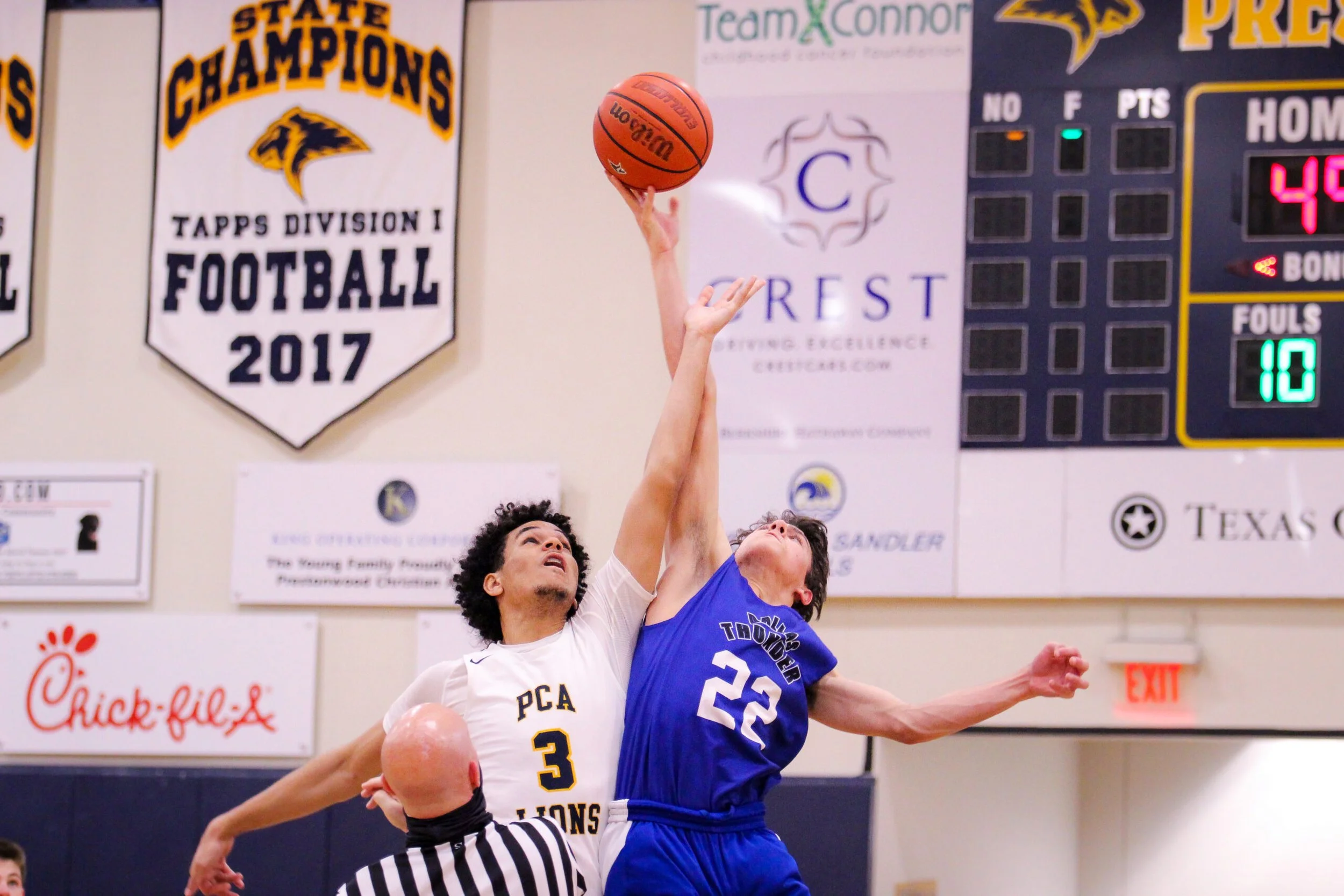 Two basketball players from opposing teams reaching for the ball during a game, with one player wearing a white jersey with the number 3 and the other wearing a blue jersey with the number 32. The background shows banners and a scoreboard.