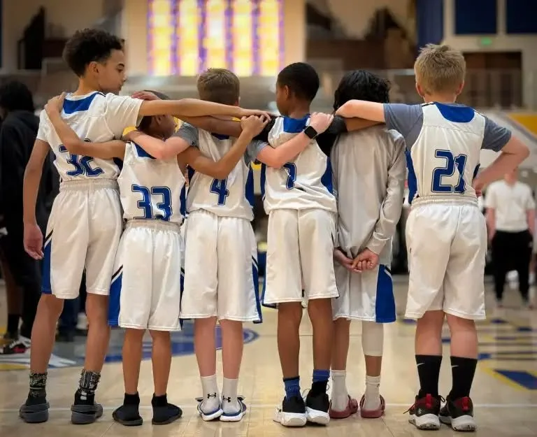 Youth basketball team huddled together on the court during a game, wearing white uniforms with blue accents.