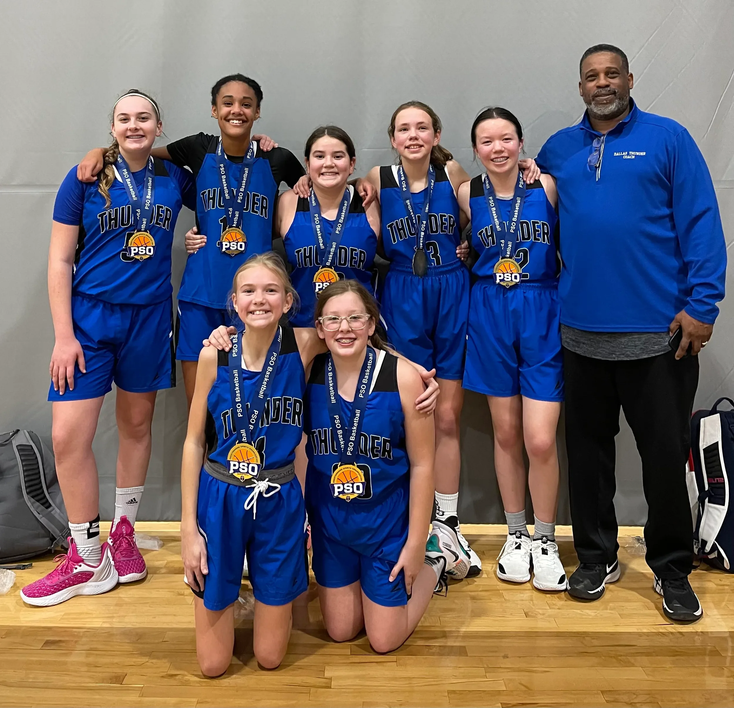 A group of five young girls and one adult man posing in a gymnasium. The girls are wearing blue basketball uniforms with 'Thunder' written on them and medals around their necks. The man is wearing a blue shirt with 'Dallas Thunder Coach' printed on it. They are smiling and standing close together, some with their arms around each other.