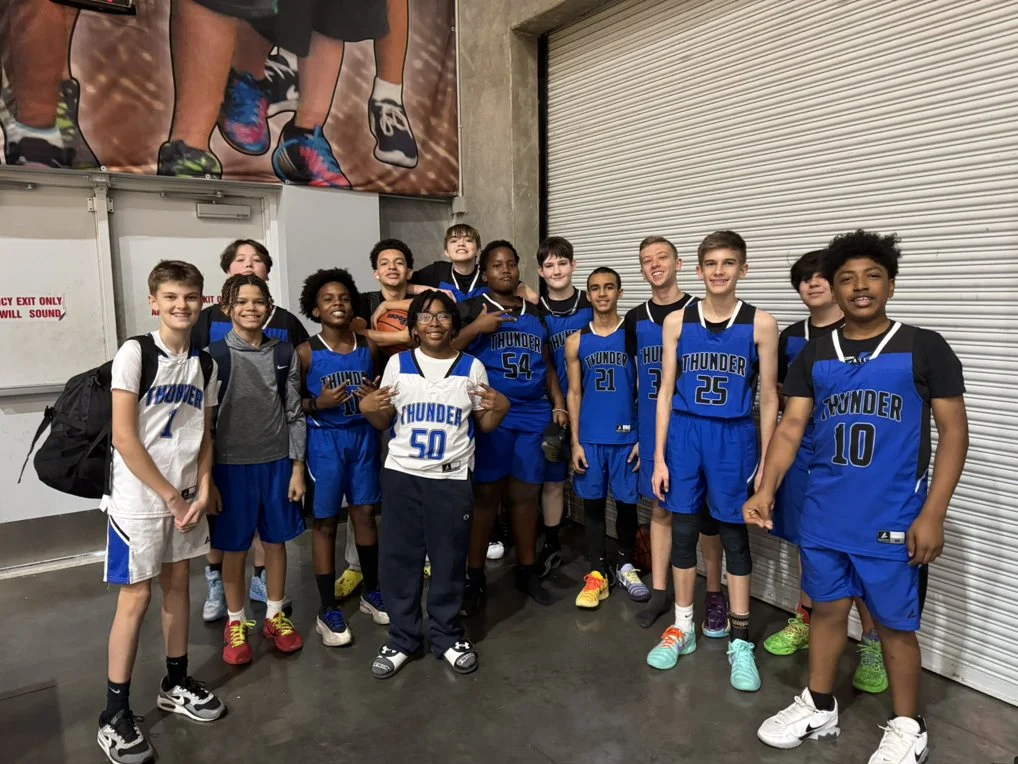 A group of young basketball players in uniforms, some in white and some in blue with 'Thunder' written on them, posing together indoors near a metal rolling door.