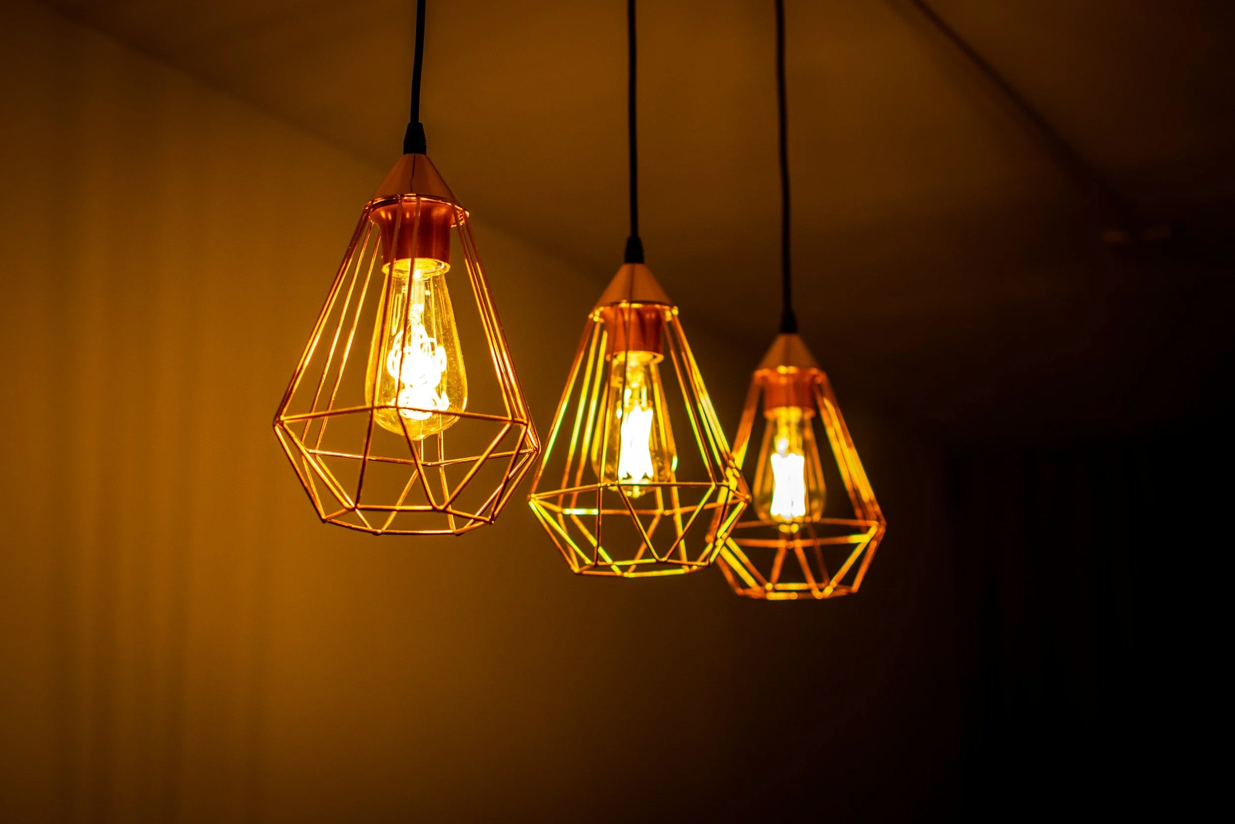 Three hanging decorative light fixtures with exposed bulbs and geometric wire frames emitting warm light in a dark room.
