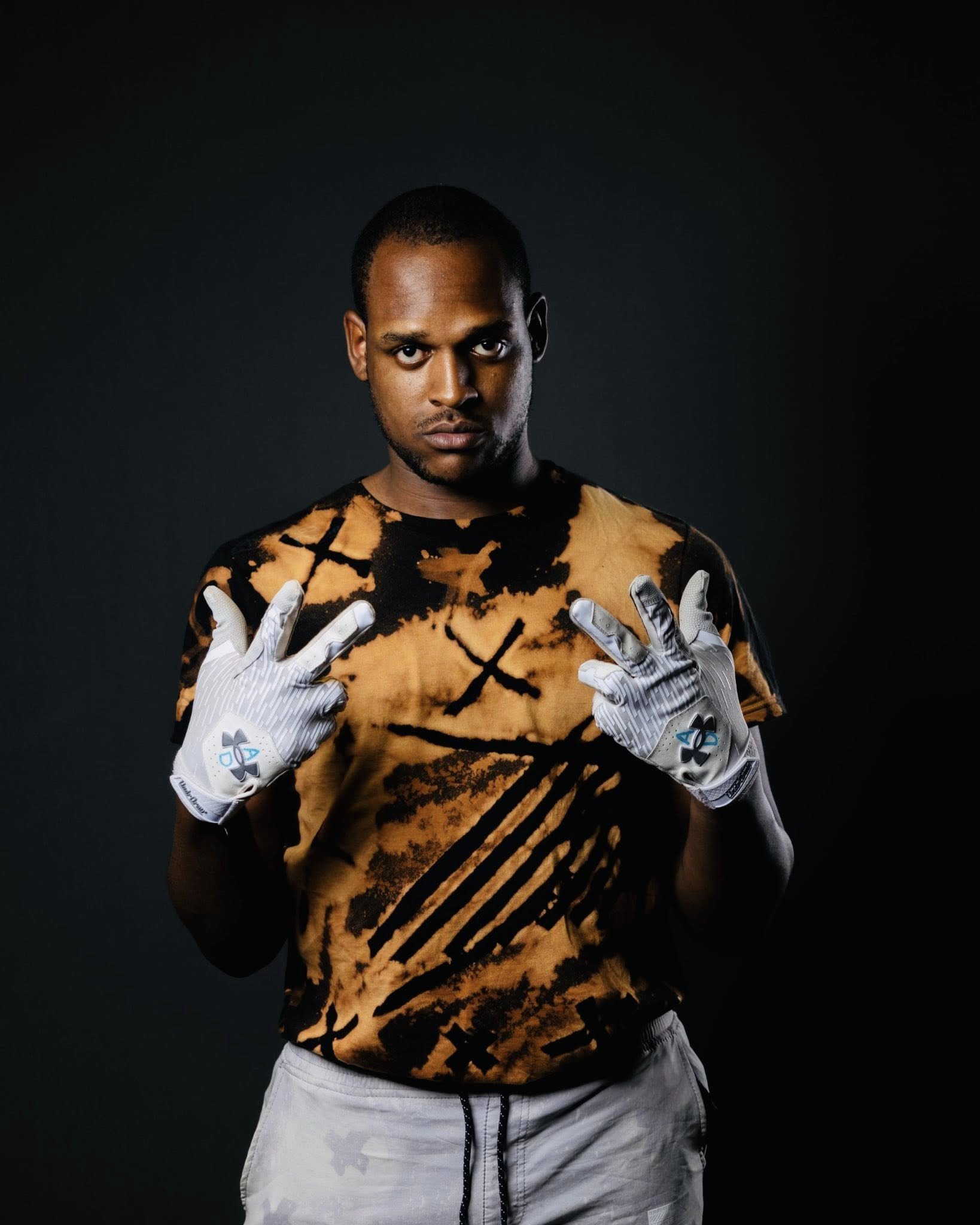 A young man with dark hair and skin is wearing a black and orange tiger-patterned shirt and white athletic shorts. He has gray and white sports gloves and is posed with his hands raised.