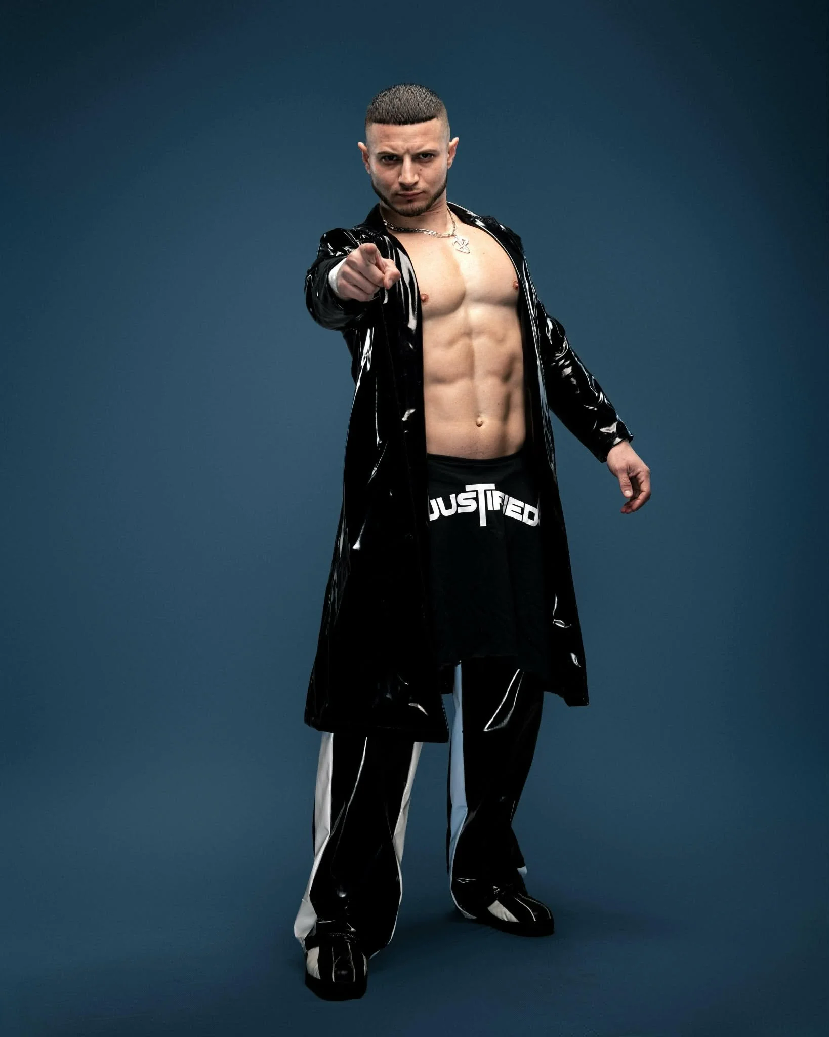 A man with short dark hair and a beard points directly at the camera. He is shirtless, wearing a long black, shiny coat with white stripes and baggy black pants with white stripes, standing against a plain blue background.