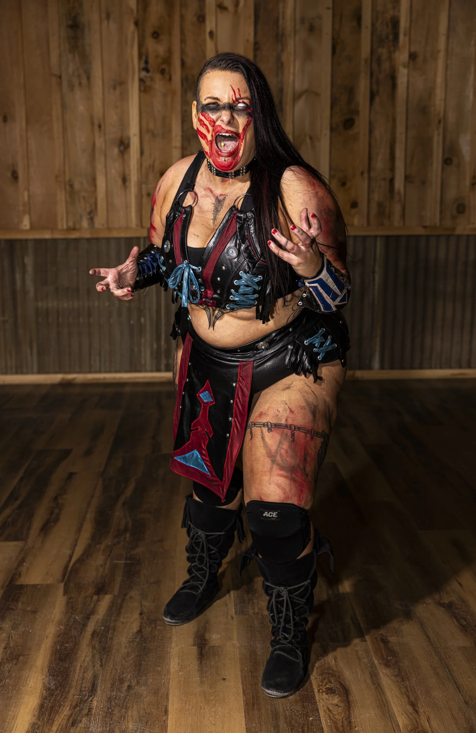 Woman with face paint, tattoos, and combat-style outfit in a wooden room.