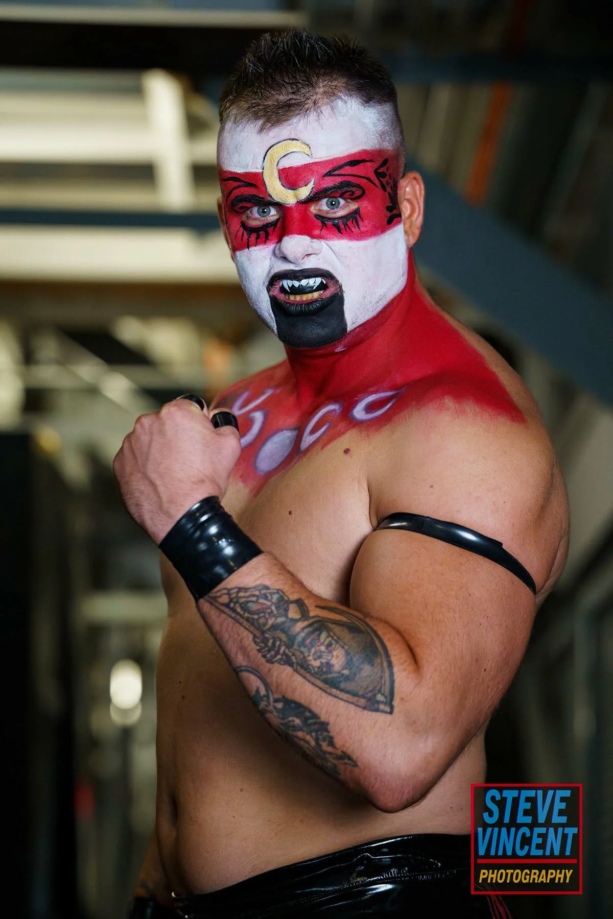 A muscular man with face paint resembling a circus clown or demon, showing teeth with vampire-like fangs, flexing his arm with a tattooed sleeve. The face paint is red, white, black, with a crescent moon shape near his forehead, and he is wearing black wristbands. The background appears to be an industrial or gym setting. The photo is signed by Steve Vincent Photography.