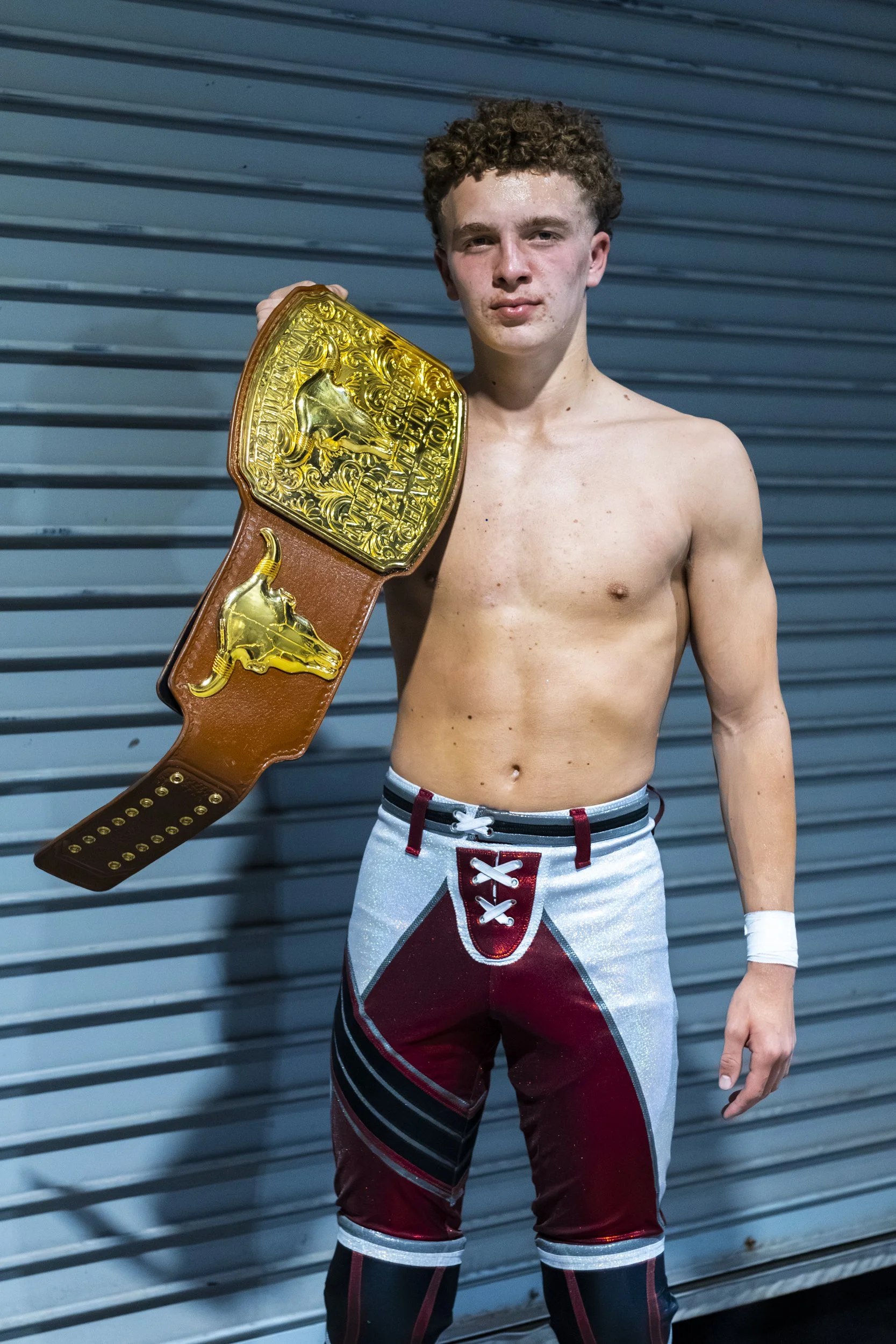 A shirtless young man with curly hair holding a large championship belt. He is wearing colorful wrestling tights with a lace-up front. He stands in front of a blue metal background.