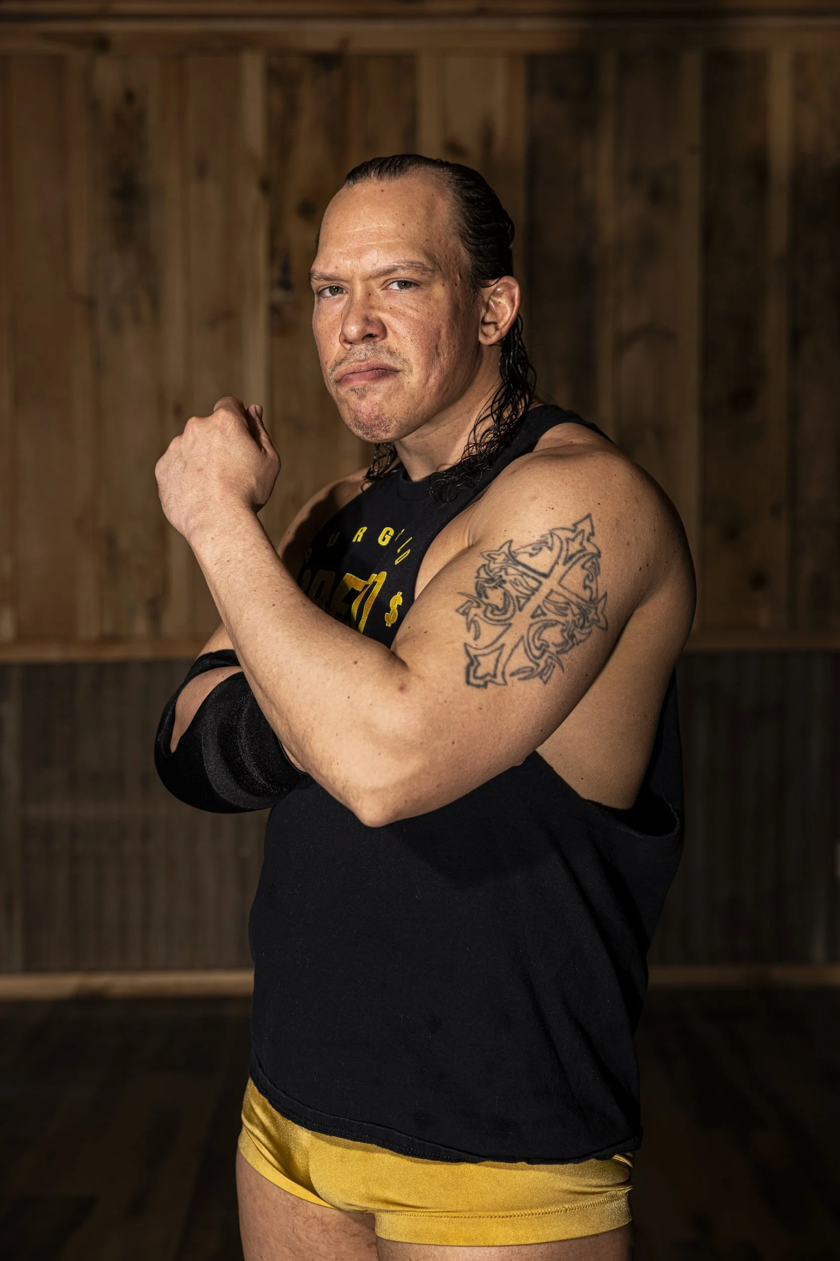 A muscular man with slicked-back hair and a serious expression, flexing his right arm, showing a tattoo on his bicep, in front of a wooden wall background.