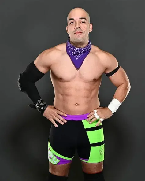 A shirtless man with a purple bandana around his neck, wearing black and neon green shorts with purple accents, a black glove on his left hand, a white wristband on his right wrist, and a black armband on his left arm, standing against a gray background.