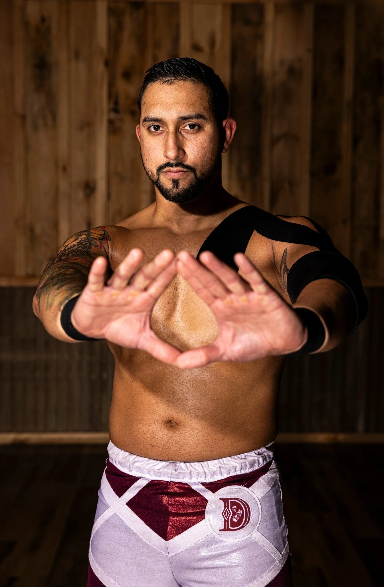 A shirtless man with tattoos on his arms and a beard, wearing shorts, standing in a wooden room, facing forward with his hands forming a diamond shape in front of him.