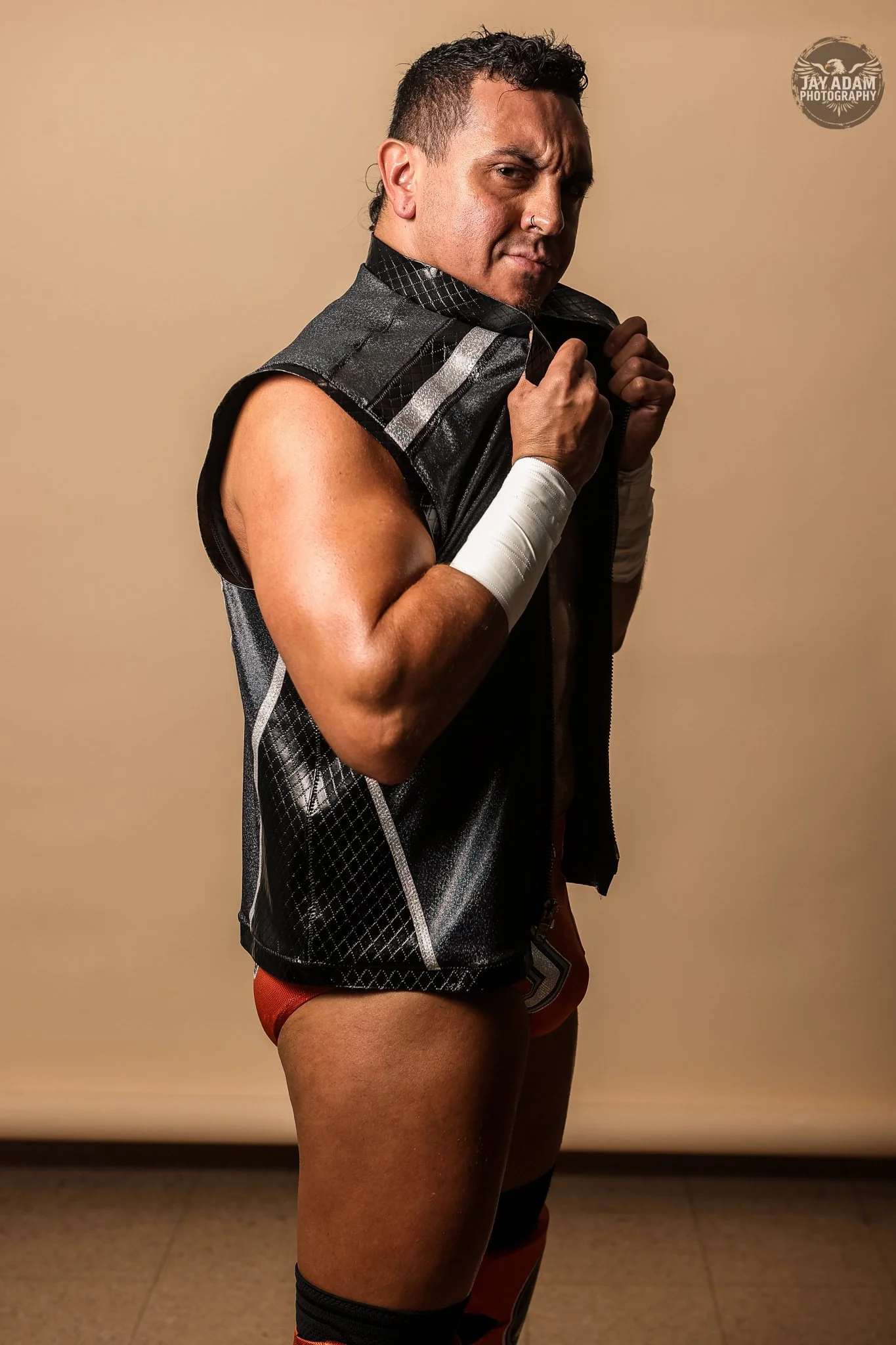 A man wearing a black athletic vest with silver accents, orange shorts, and black knee-high socks poses with a confident expression, holding the collar of his vest with both hands, against a plain beige background.