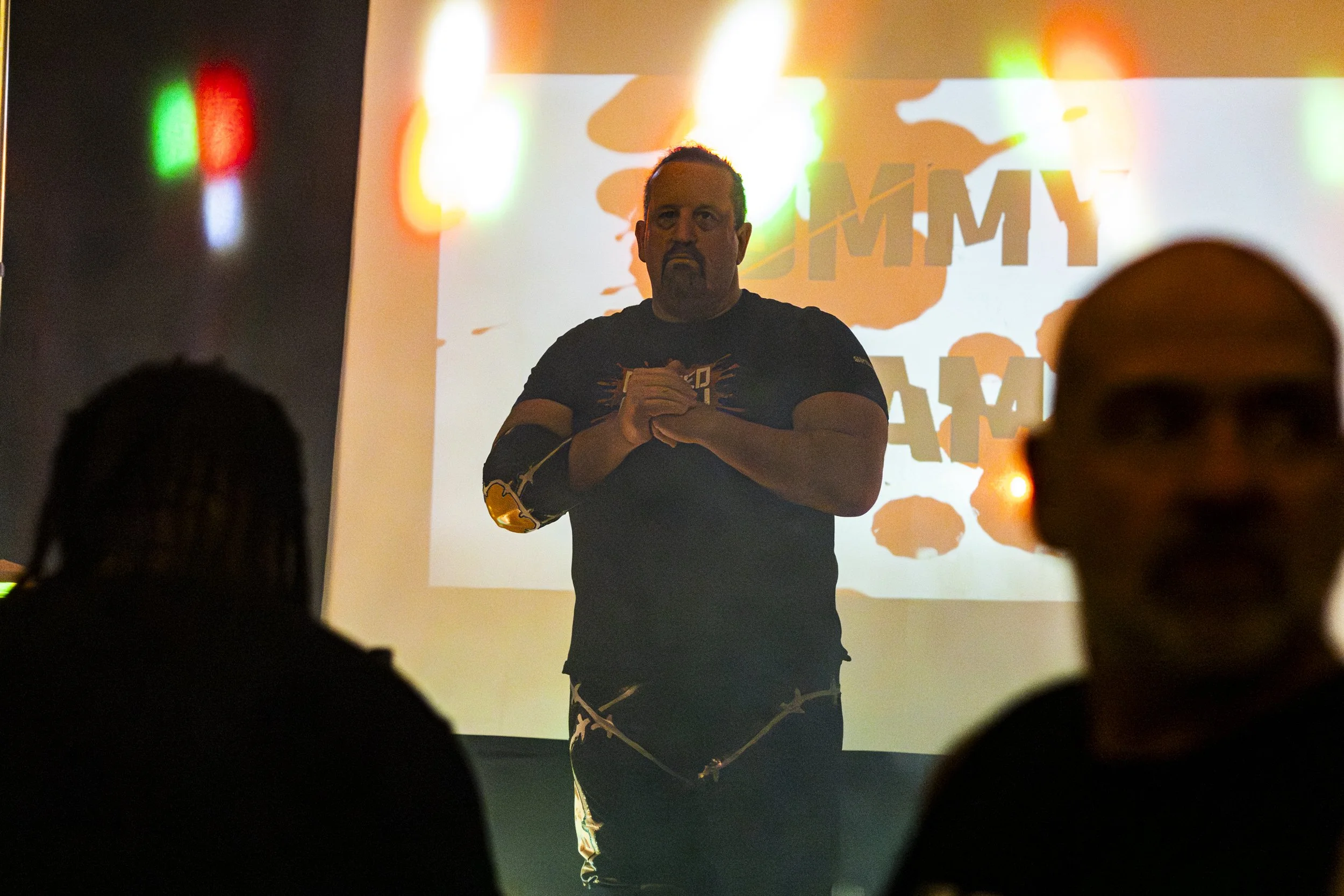 A man with a beard and mustache stands on stage in front of a screen with an orange and white background. He has a serious expression, with hands clasped in front of him. Audience silhouettes are visible in the foreground, and colorful lights are reflected on the screen.