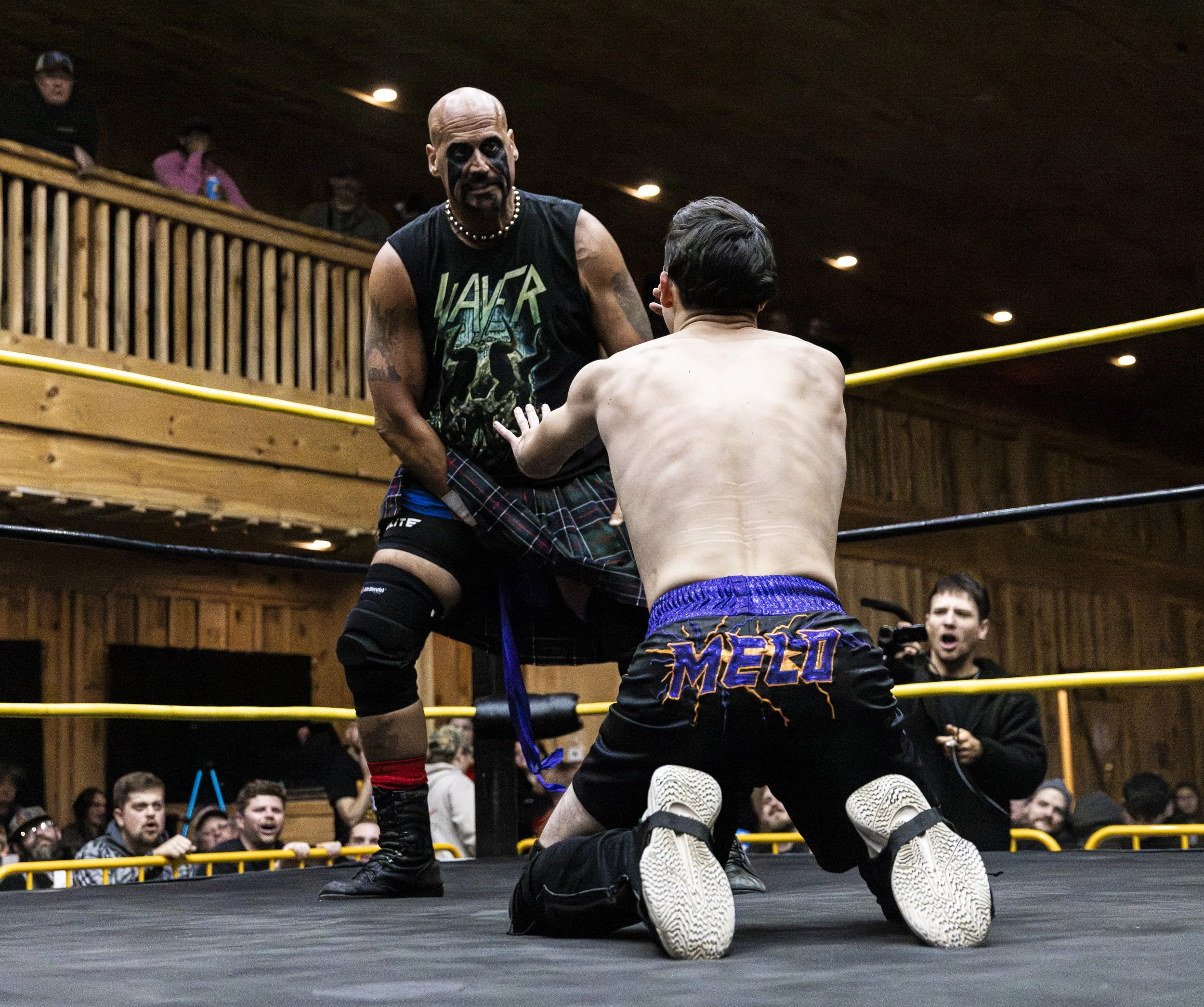Two wrestlers engaged in a wrestling match inside a ring, surrounded by an audience. One wrestler is a bald man with face paint and tattoos, wearing a sleeveless shirt, plaid shorts, and knee-high socks. The other wrestler, whose back is visible, has dark hair and is wearing black shorts with colorful lettering and design. There are spectators watching and cheering in the background.
