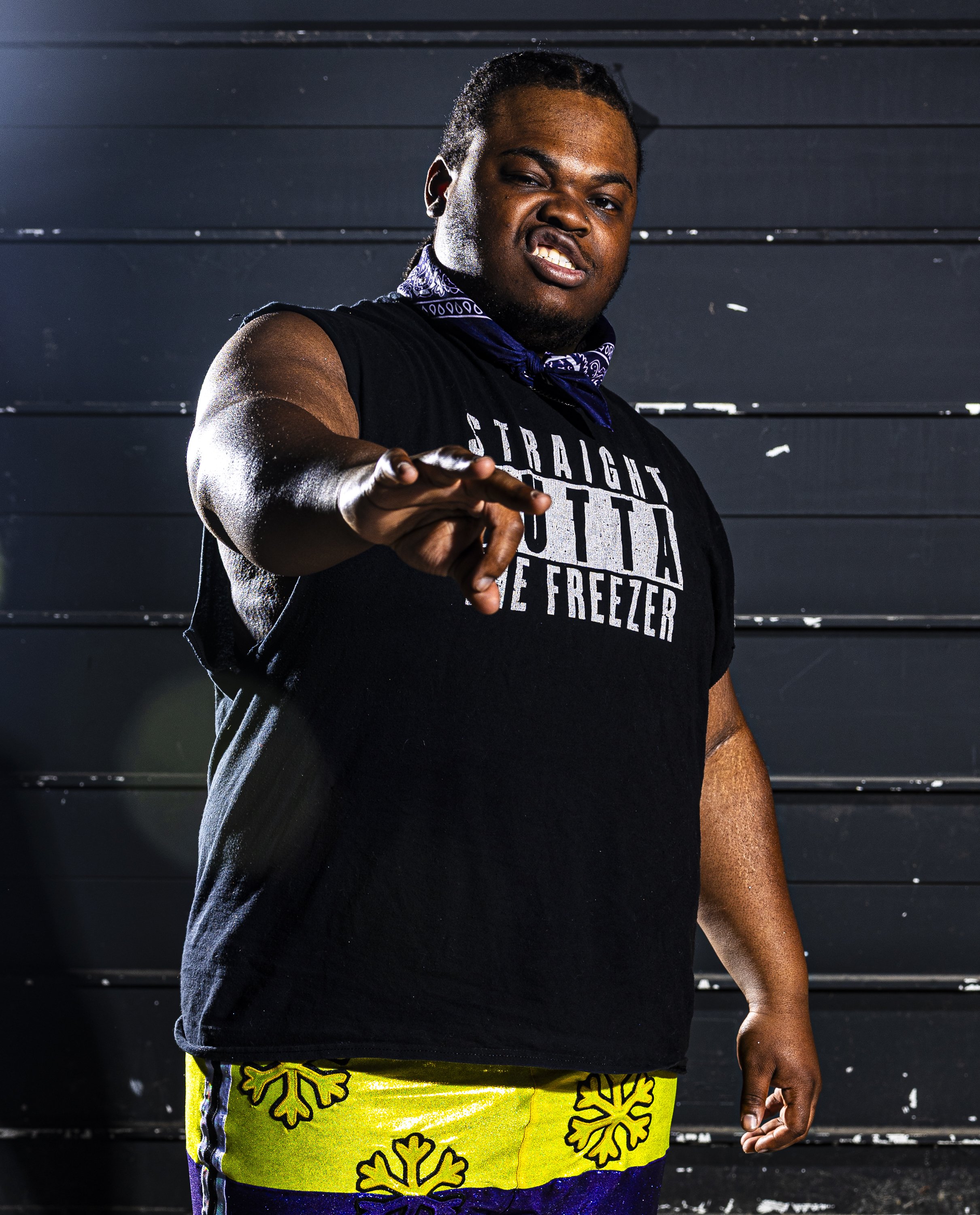 A person posing in front of a dark horizontal panel background, wearing bright yellow shorts with black and purple accents and a black T-shirt with white text, holding one arm out with fingers partially curled, and making a playful facial expression.