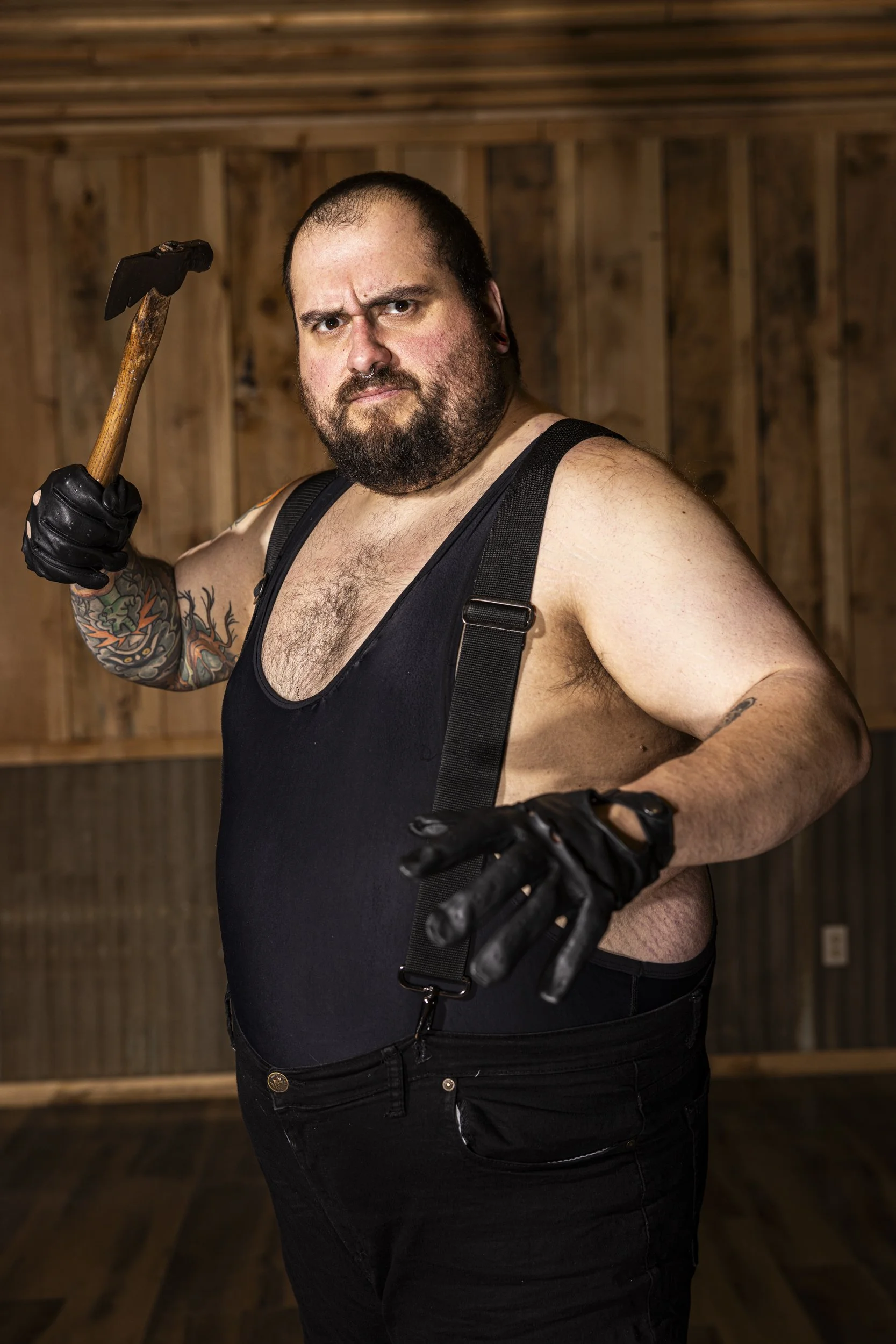 A man with a shaved head and beard, wearing a black tank top, black gloves, and suspenders, holds a hammer over his shoulder. The background features a wooden wall and floor.
