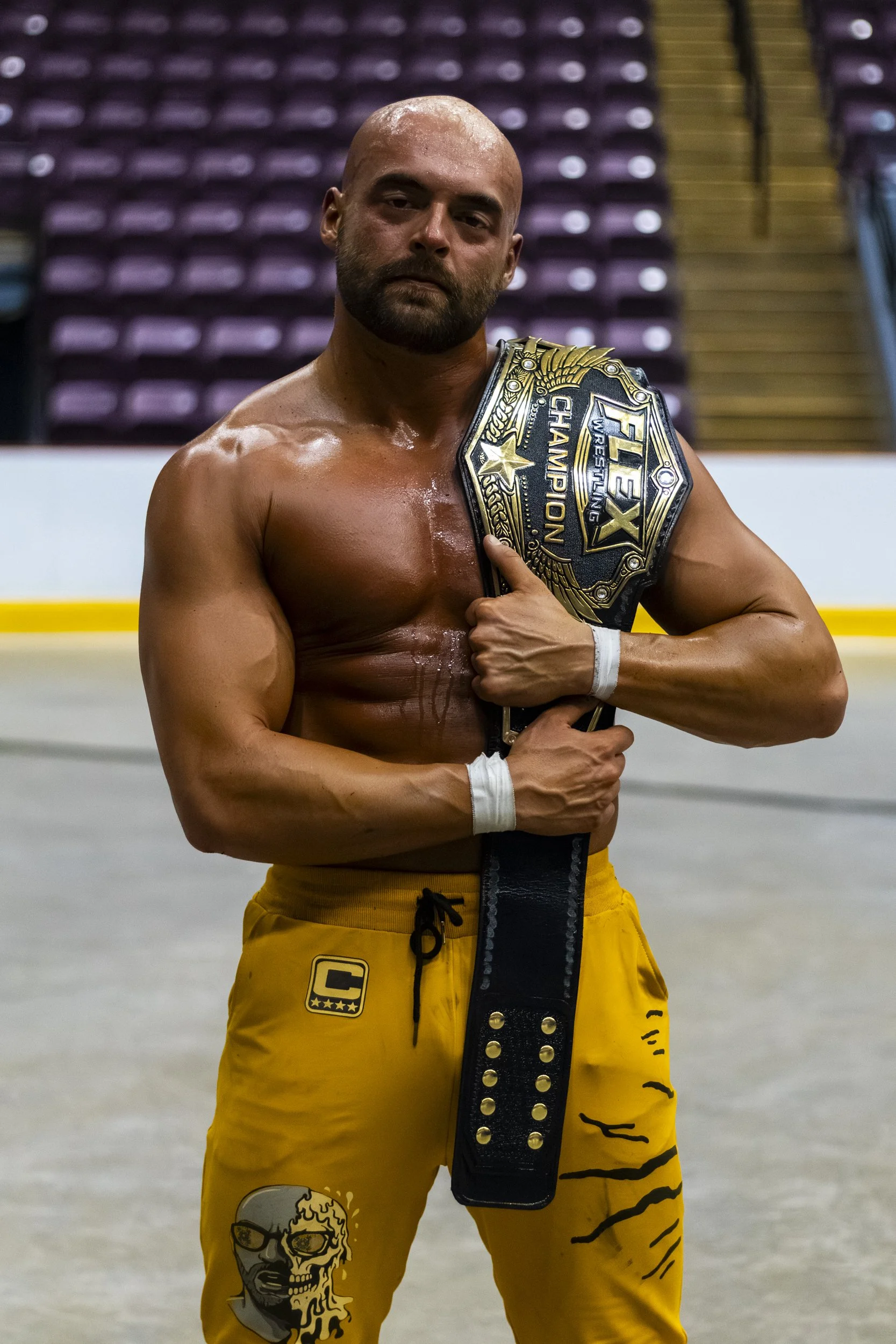 A shirtless male athlete with a beard and a shaved head holding a championship belt over his shoulder in a sports arena with empty purple seats in the background. He is wearing yellow shorts with a skull illustration and black tiger stripe patterns.