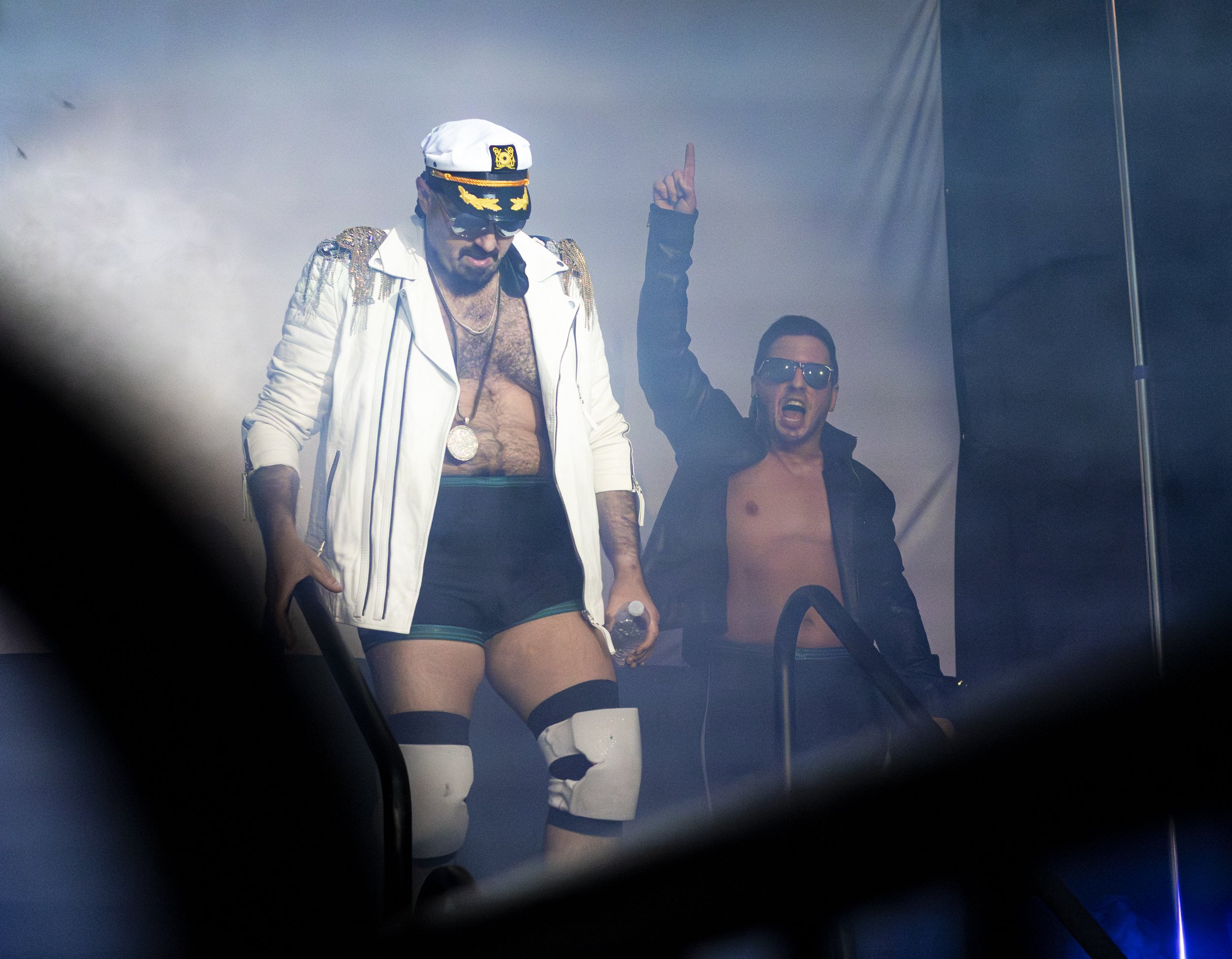 Two men on stage; one wearing a captain's hat and sunglasses, with gold jacket draped over shoulders, pointing upward; the other with shout, shirtless, wearing sunglasses, raising one arm.