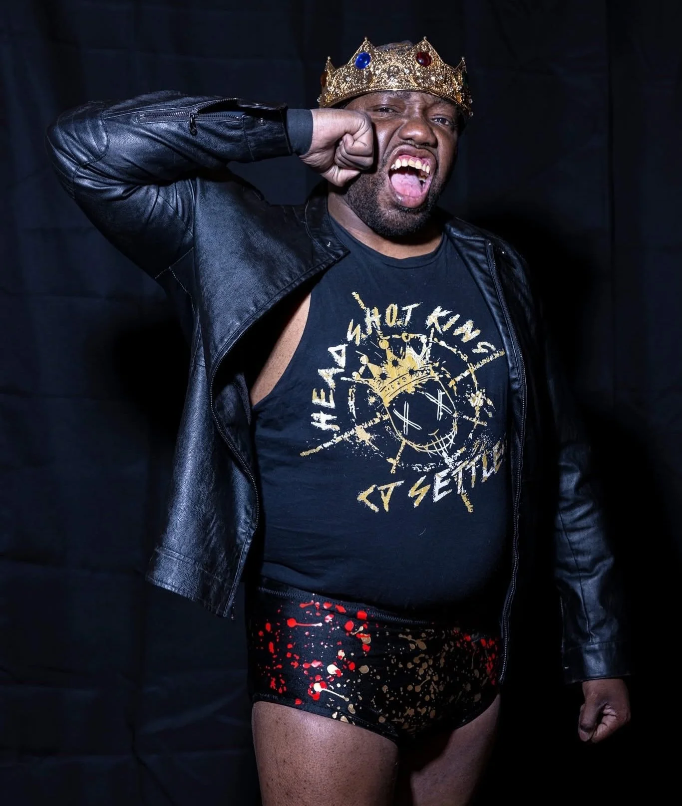 A man wearing a crown, black leather jacket, a graphic T-shirt, and colorful splatter shorts, making a fist to his face with an angry expression.