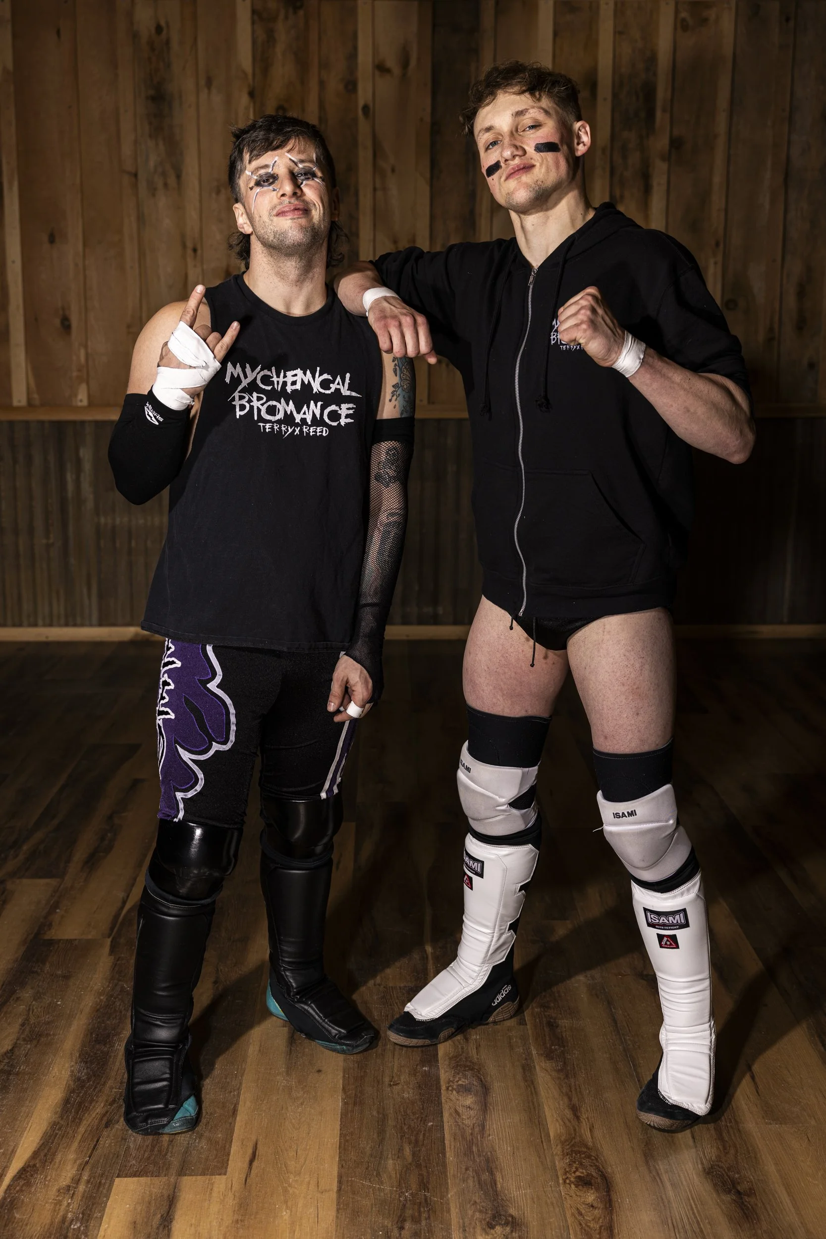 Two male wrestlers in a gym, one with face paint and the other with black face markings, both wearing wrestling gear and standing on a wooden floor with a wooden wall background.