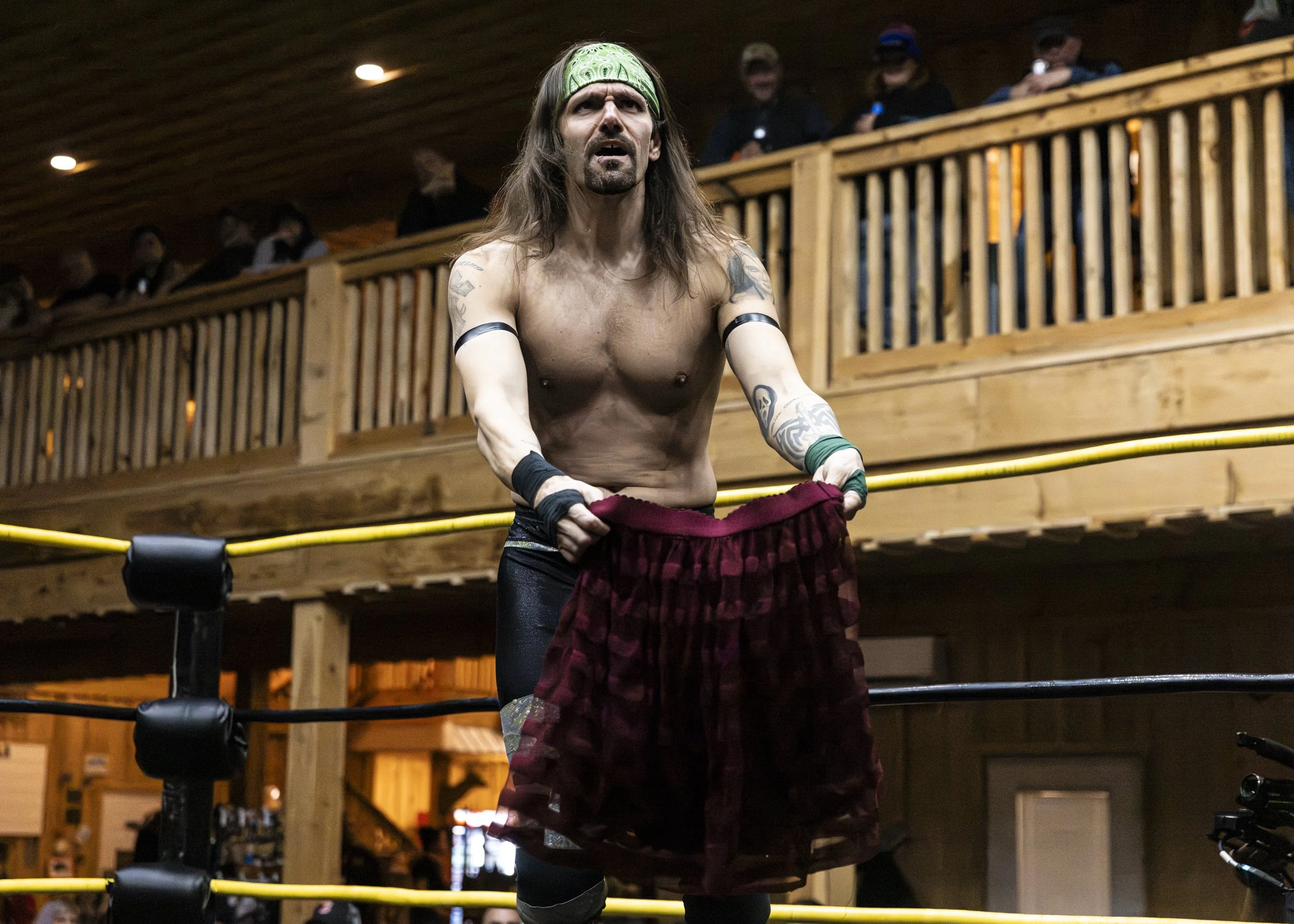 A shirtless male wrestler with long hair and tattoos, wearing a green headband, standing inside a wrestling ring.
