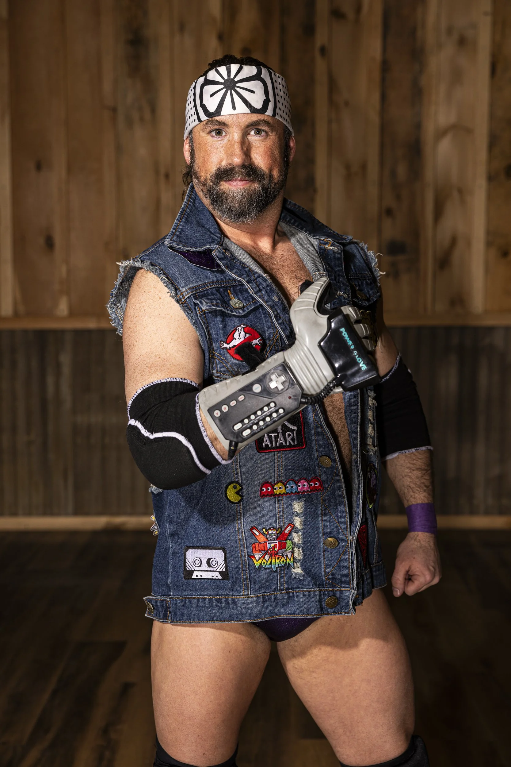A man dressed as a 1980s arcade character with a denim vest decorated with various patches and pins, including Pac-Man and Transformers patches, wearing a bandana, fingerless gloves, and knee-high socks, standing in front of a wooden wall.