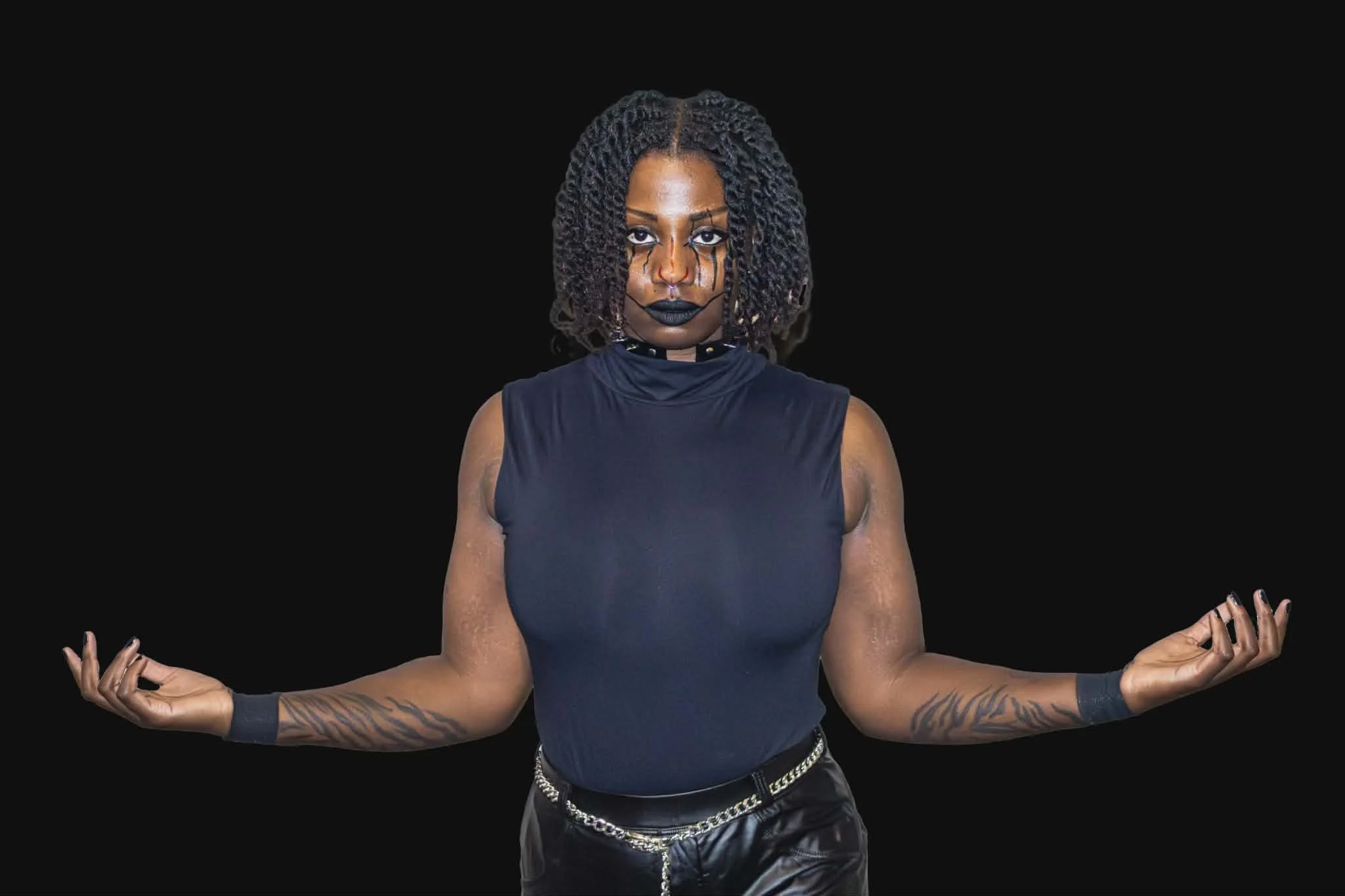 A woman with curly dark hair in a meditation pose, wearing a sleeveless black shirt and black gloves, with tattoos on her forearms, standing against a black background.