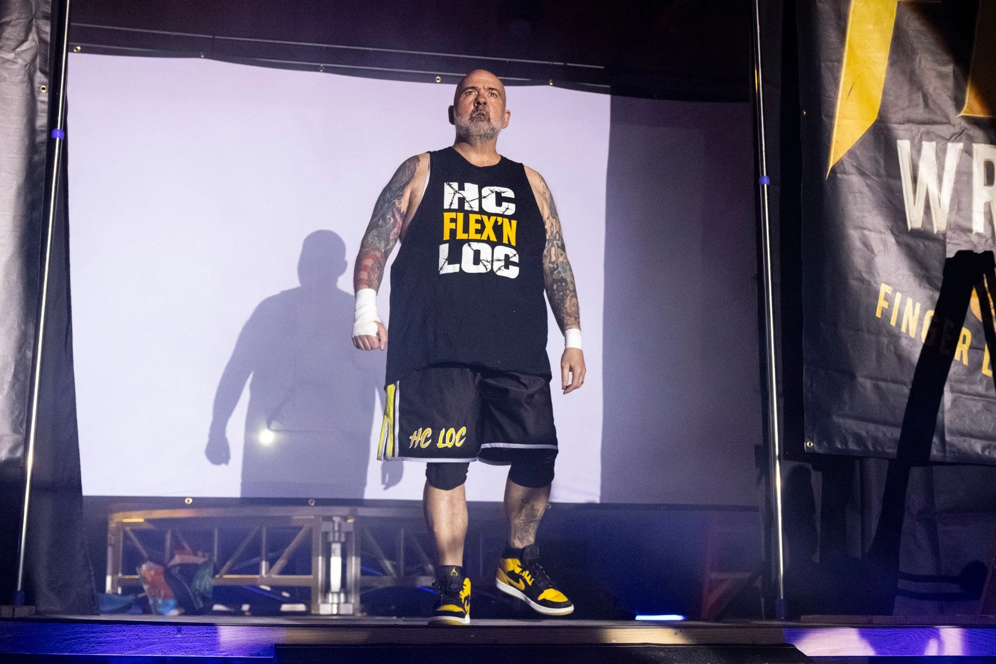 A muscular man with tattoos on both arms, dressed in a black tank top and matching shorts, walking out on a stage. The tank top and shorts feature the text 'HC LOC' and 'FLEX'N'. He has a bald head and a grey beard, and is wearing black and yellow sneakers. The stage backdrop shows the event name 'WRESTLE' partially visible on a black and yellow banner.