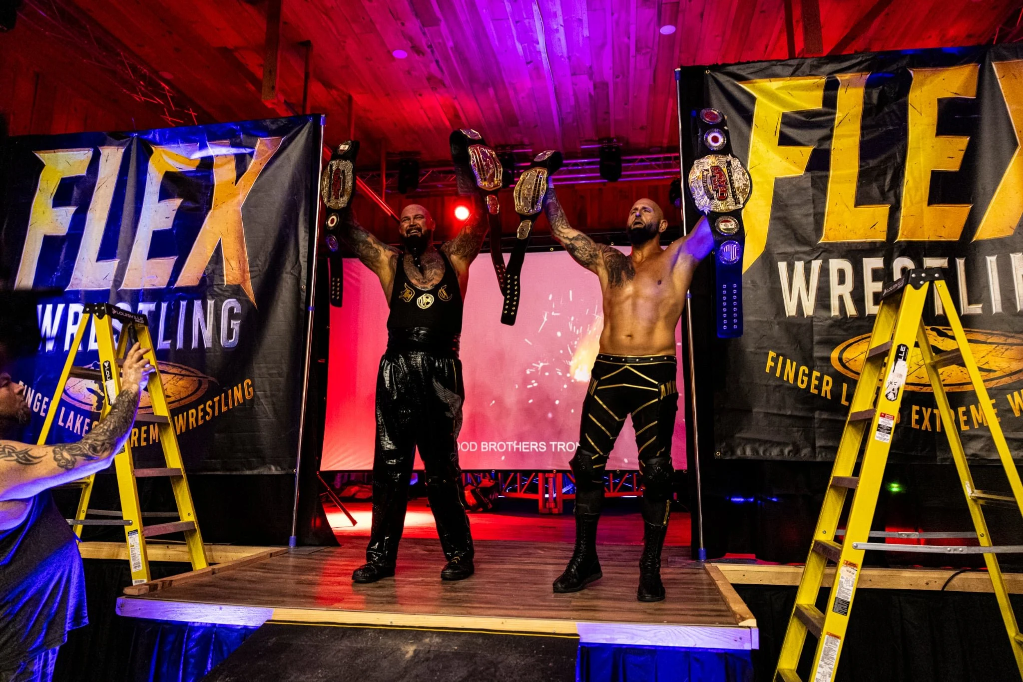 Two male wrestlers are celebrating with belts raised in front of an audience at a wrestling event. They are standing on a small stage with yellow ladders on either side, covered by banners reading 'FURY' and 'WRESTLING.' The setting has colorful lighting, and a large screen behind them shows the event's name.