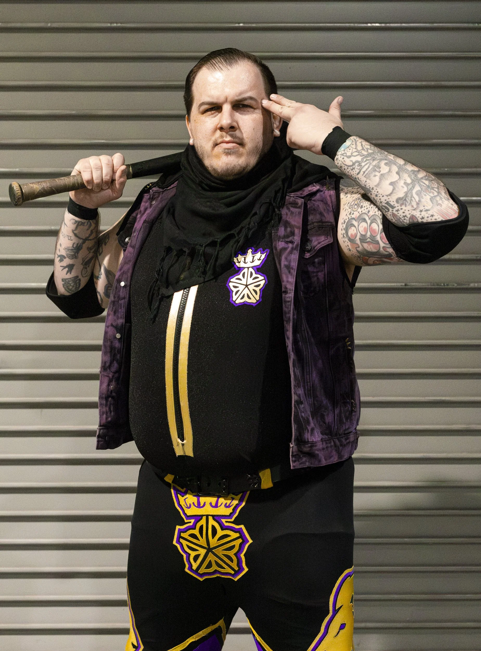 A man with tattoos on his arms is standing in front of a metal roll-up door. He is holding a baseball bat over his shoulder and is making a gesture with his right hand near his head. He is wearing a black shirt with a colorful logo, a purple and black vest, and black shorts with purple and yellow designs.