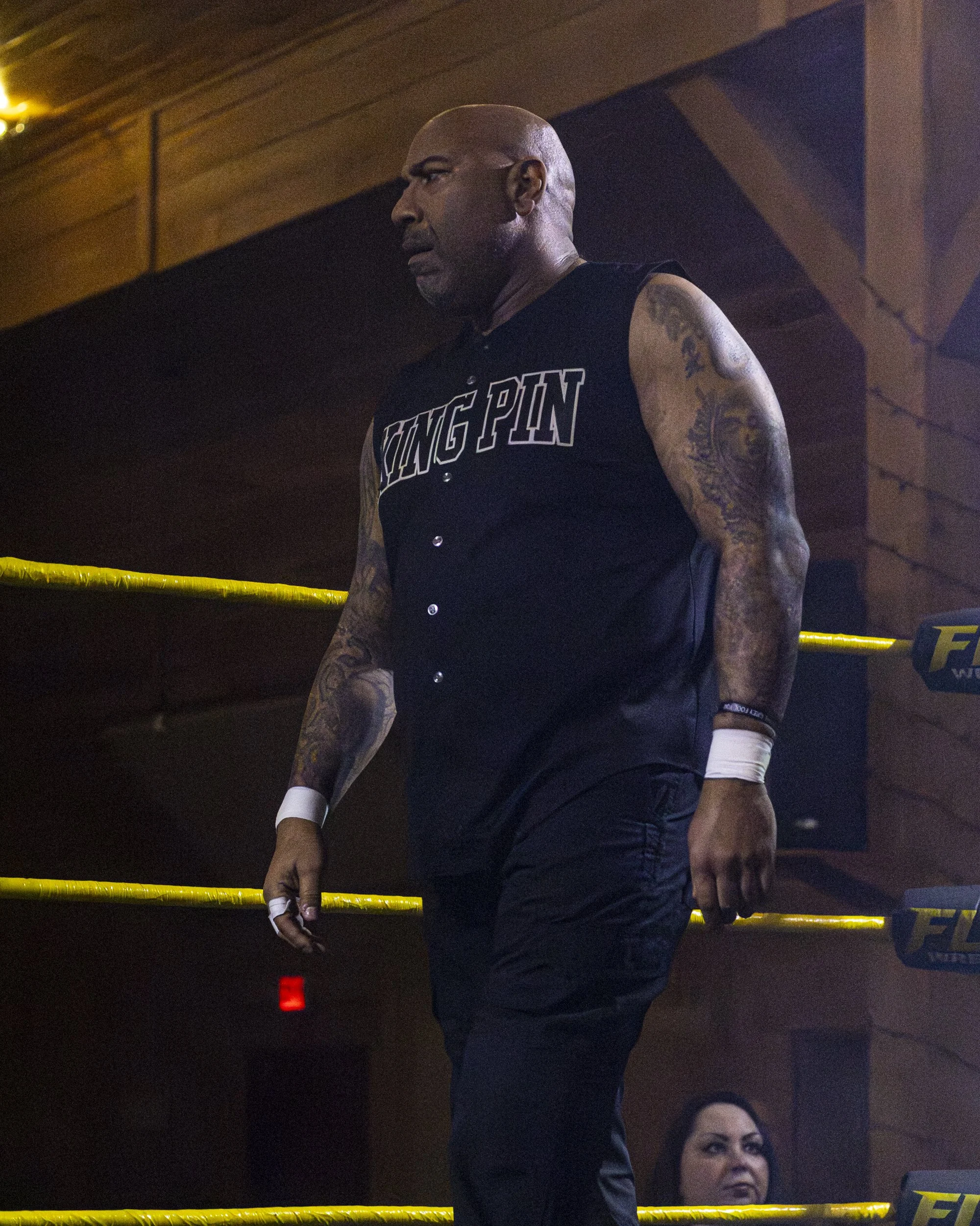 A bald man with tattoos on his arms wearing a sleeveless black shirt with 'KING PIN' written on it, standing inside a wrestling ring with yellow ropes, under dim lighting, a woman is partially visible in the background.