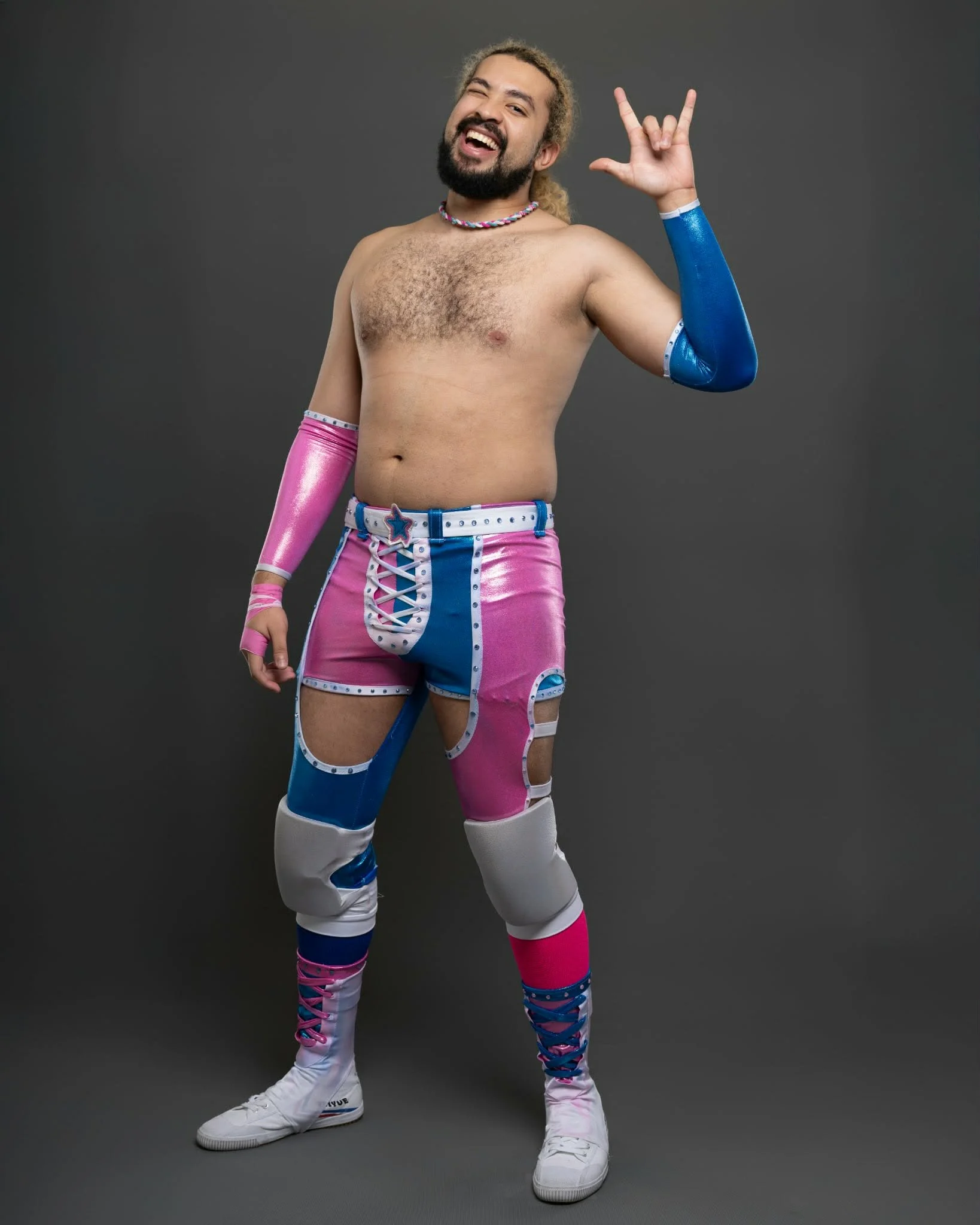 A man with long curly hair, a beard, and light skin is shirtless and making a rock sign with his right hand. He is smiling, winking, and showing his teeth. He is wearing colorful, shiny, pink, blue, and white pants with a star on the belt and white sneakers with high socks. One arm is covered with a shiny blue sleeve, and the other arm has a pink sleeve. He is standing against a plain gray backdrop.