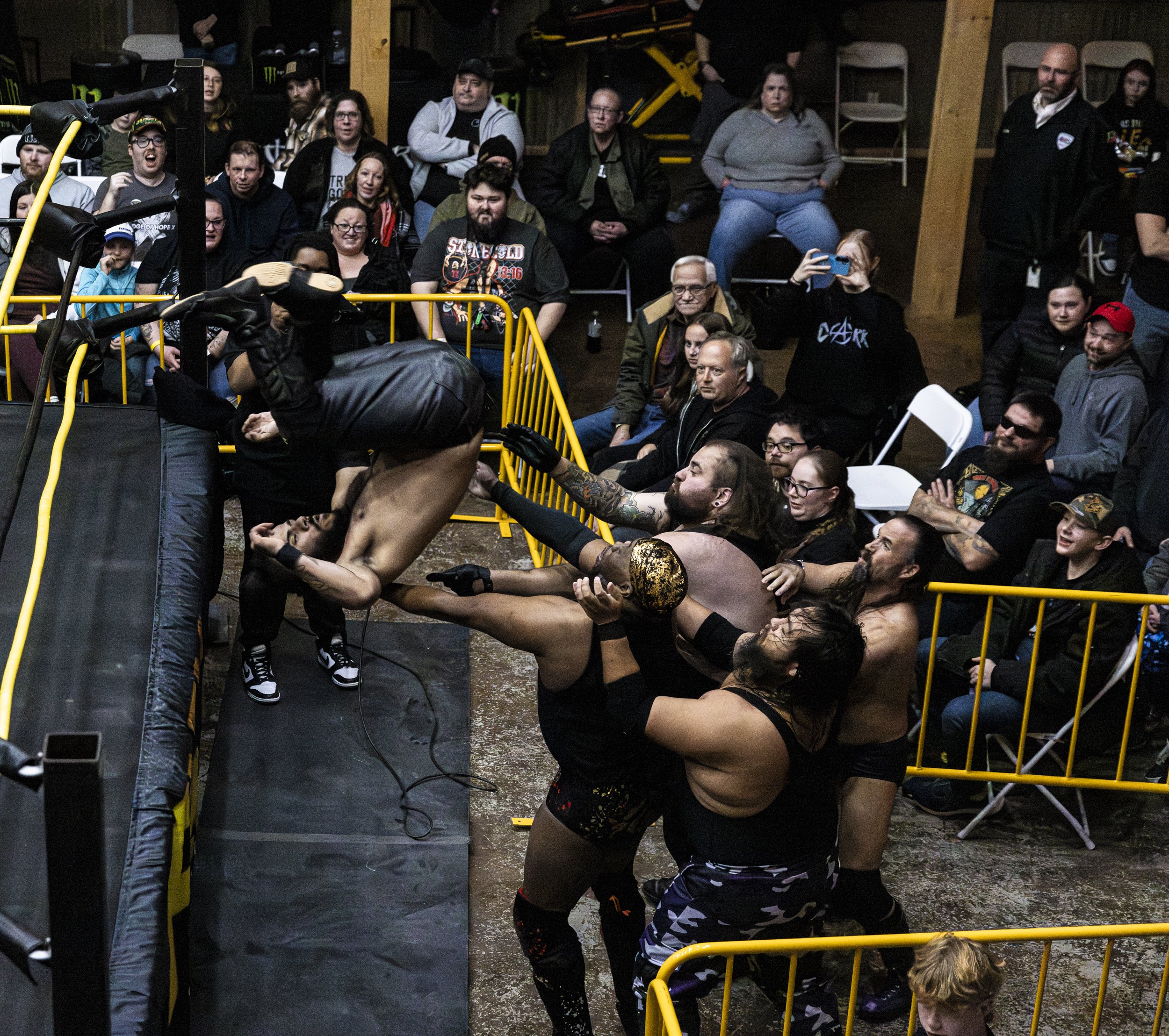 A crowd of people sitting and standing in a dark indoor space watching a wrestling match. Some are taking photos or videos, and a wrestler is in action near the ring with ropes and yellow barriers.