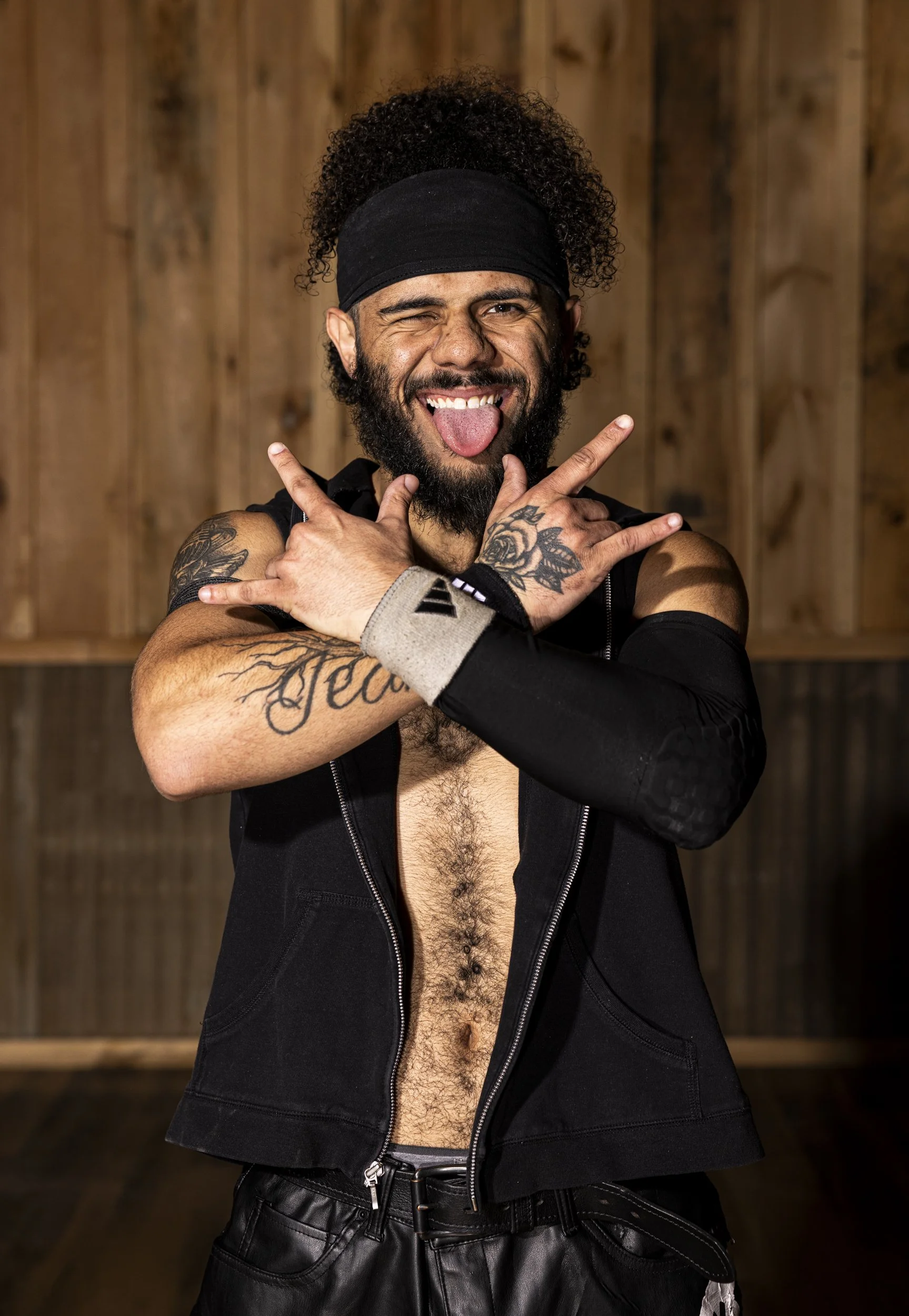 A man with a beard and curly hair wearing a black headband, black sleeveless vest, and black leather pants, making a playful pose with tongue out and fingers pointing in the air, standing against a wooden background.