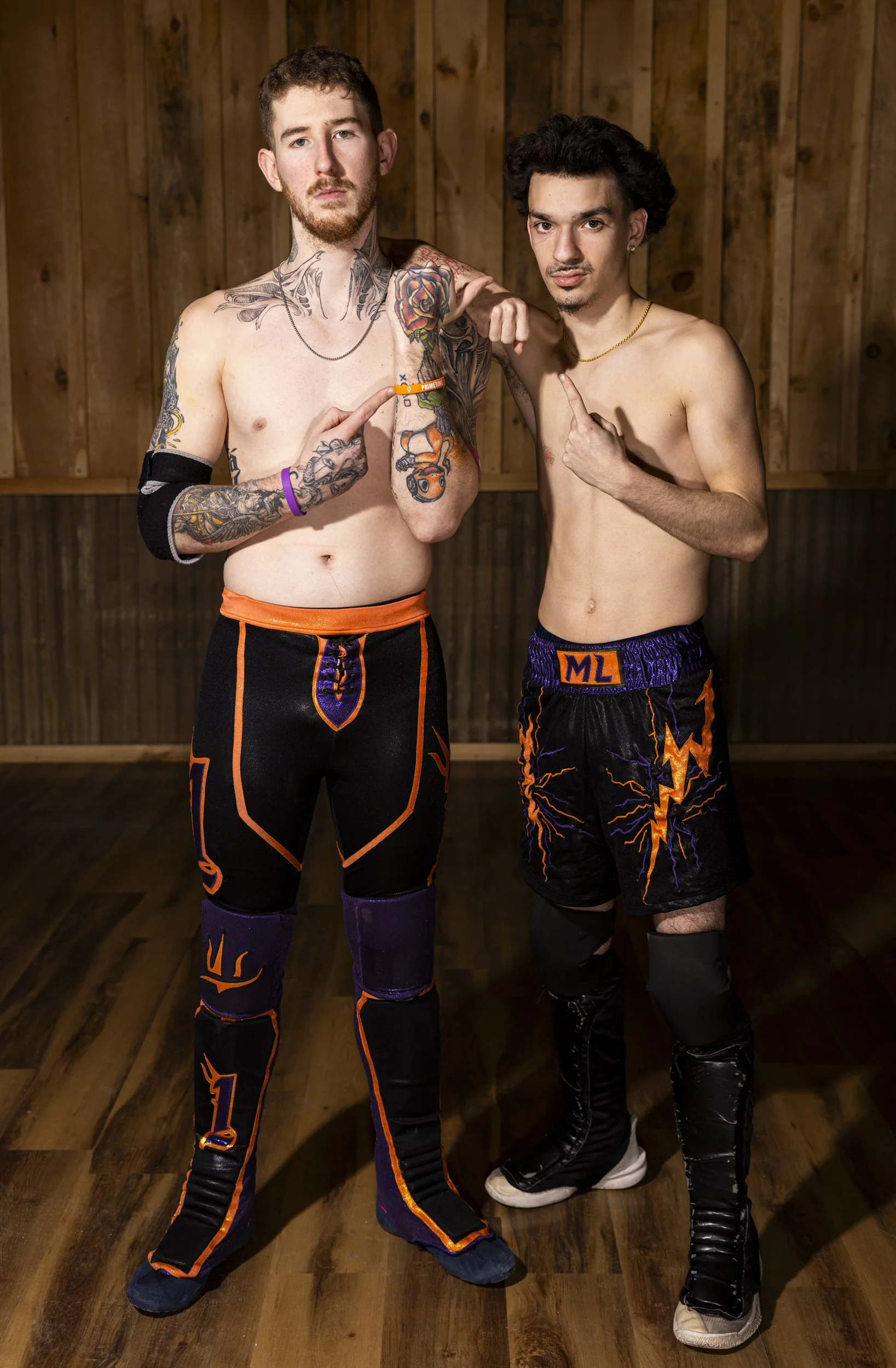 Two shirtless male wrestlers standing in a wrestling ring with a wooden wall background, wearing black and orange wrestling gear, pointing at each other.