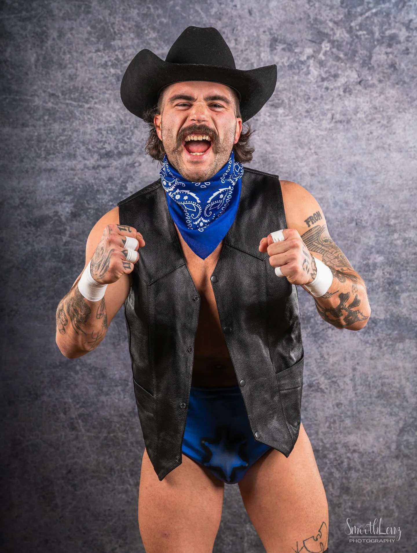 A man wearing a black cowboy hat, blue bandana, black leather vest, and blue underwear, posing with fists raised. He has tattoos on his arms and legs, facial hair, and is against a gray textured background.