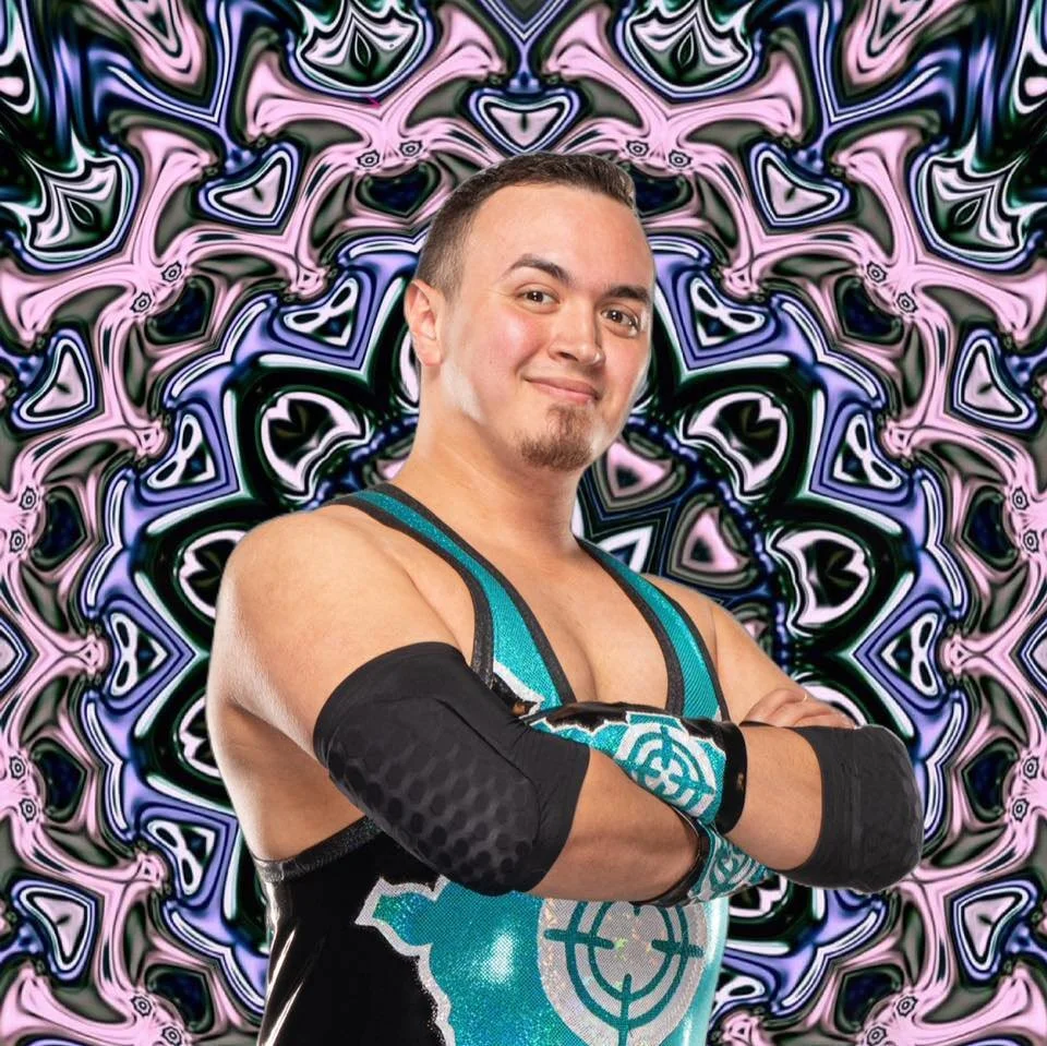 A man with short dark hair, a goatee, and a confident smile, wearing a wrestling singlet and arm sleeves, standing with his arms crossed in front of a colorful, abstract psychedelic background.