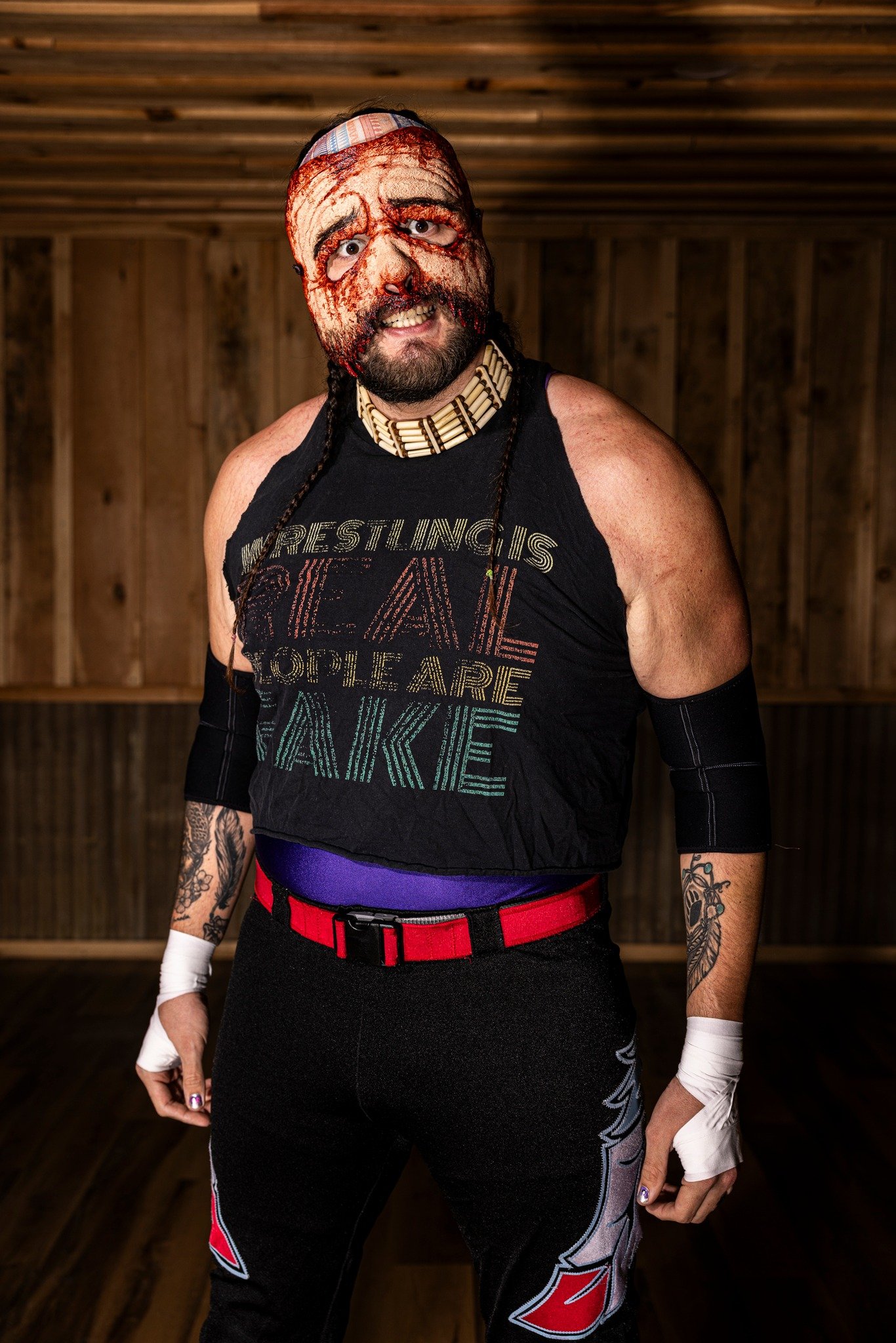 A man wearing wrestling gear with a mask painted on his face, a choker, and tattoos on his arms, standing inside a wooden gym or training room.